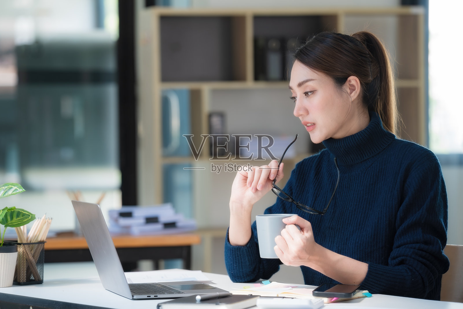 一位亚洲女商人一边用笔记本电脑工作，一边拿着一杯热咖啡。照片摄影图片