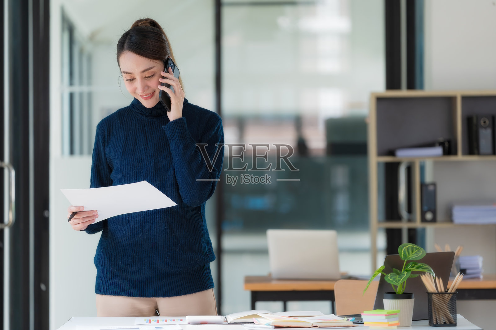 一位亚洲女商人正在办公室里工作，她手里拿着一份文件报告，并使用手机。照片摄影图片