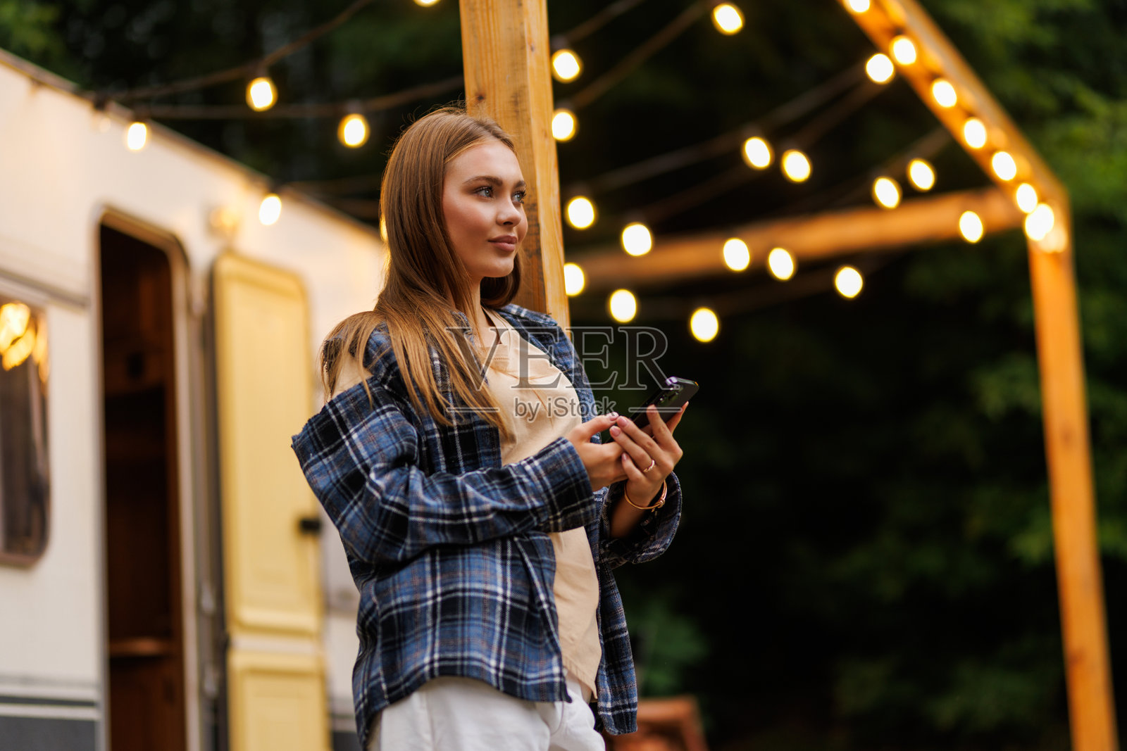 夜晚，一位快乐的女士在户外露营车旁，一边放松一边浏览手机，脸上带着笑容。照片摄影图片