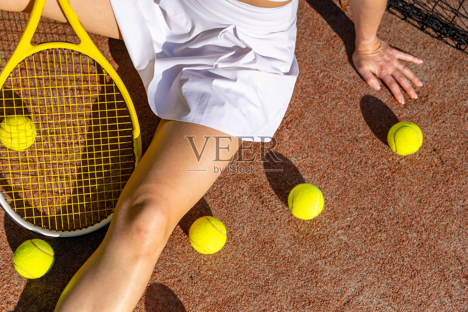 阳光下，女网球运动员坐在球场上休息，身旁放着网球拍和散落的网球。照片摄影图片