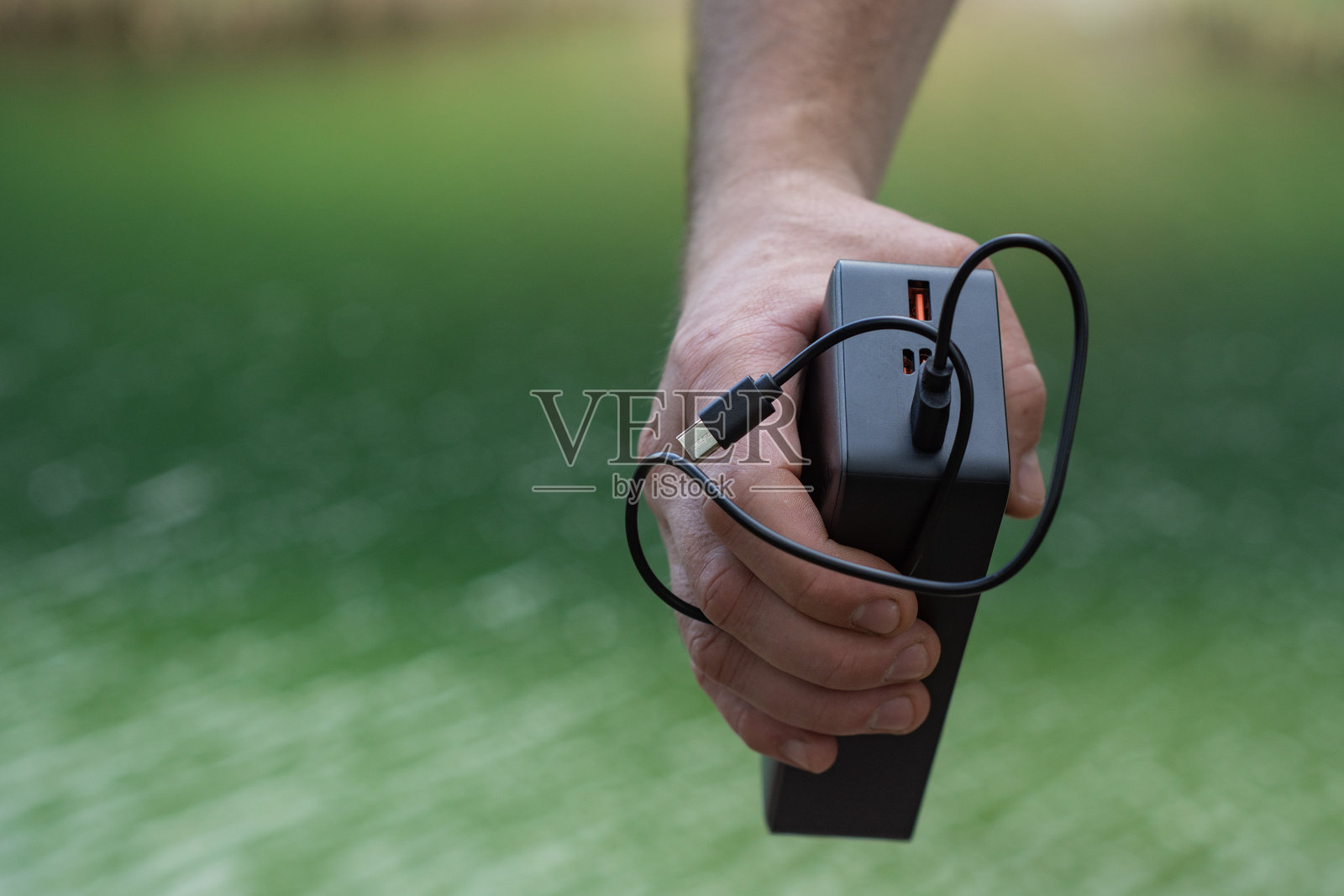 手持黑色便携式充电宝，背景是模糊的、充满活力的绿色水景。代表移动充电、便携式技术和度假时保持联系的概念。照片摄影图片