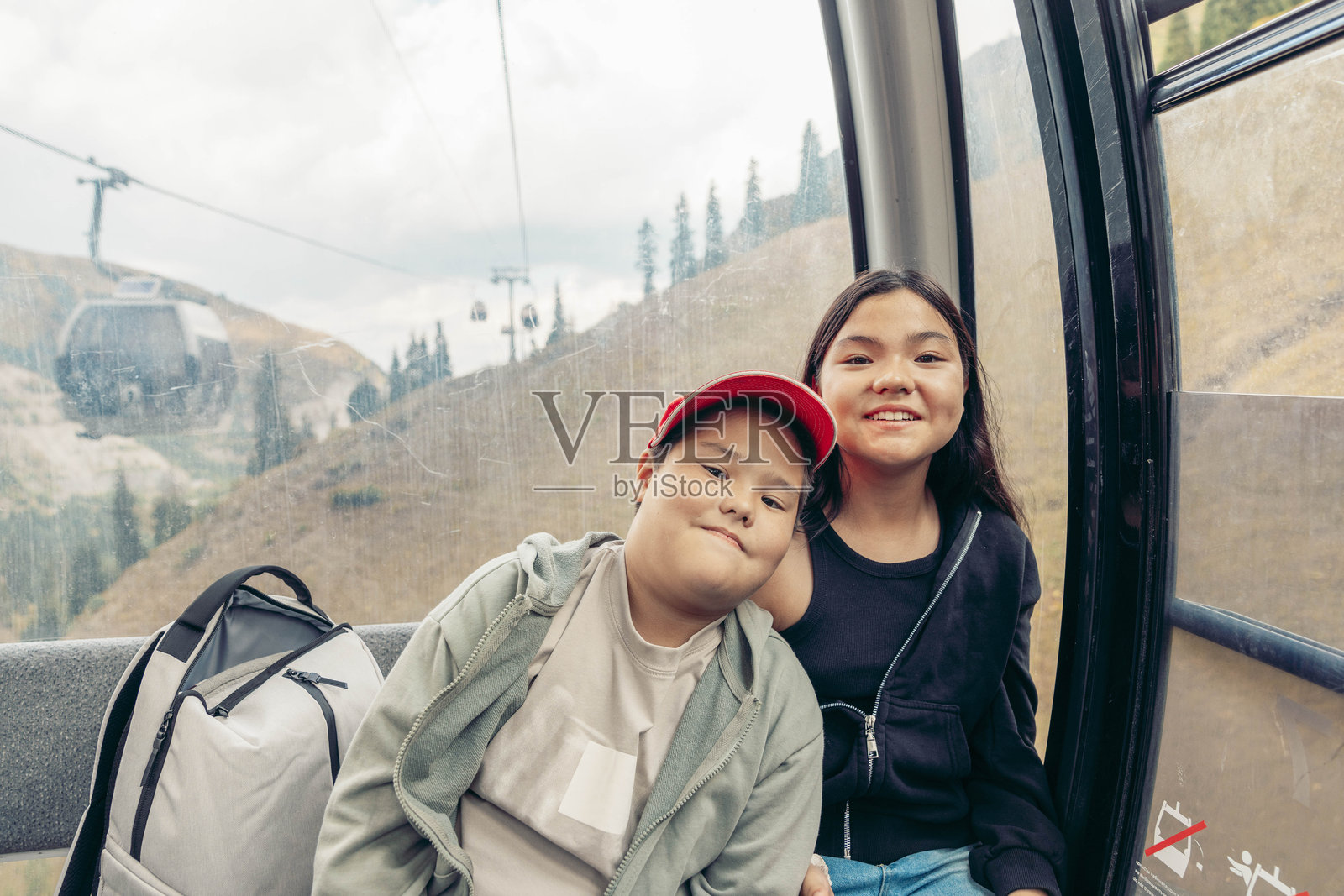 两个孩子在山里坐缆车。一个戴红帽的亚洲小男孩和一个亚洲小女孩一起微笑。背景是风景优美的山景。照片摄影图片
