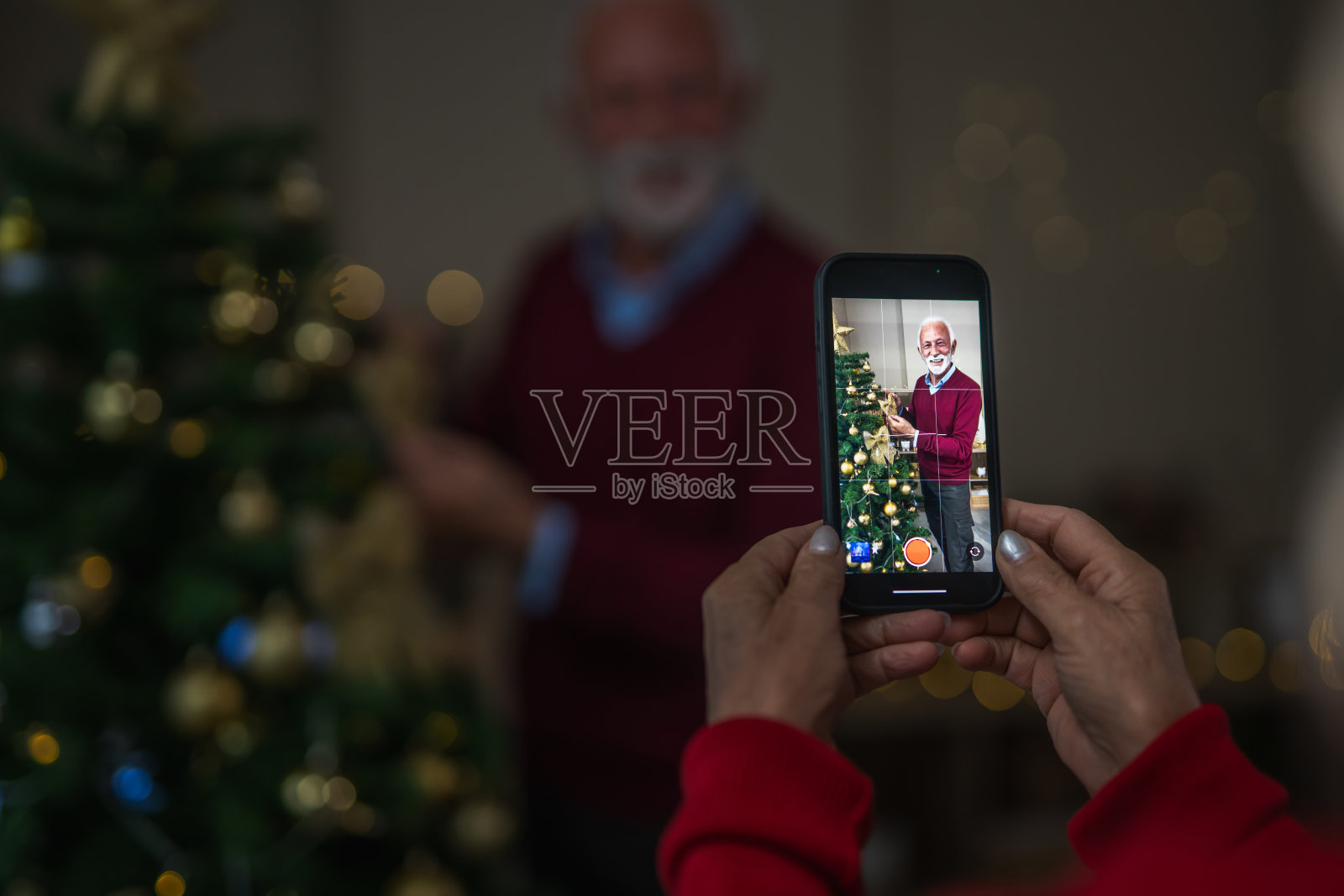 一位年长的女性用智能手机拍摄她的丈夫装饰圣诞树。/ A senior woman films her husband decorating a Christmas tree on a smartphone.照片摄影图片