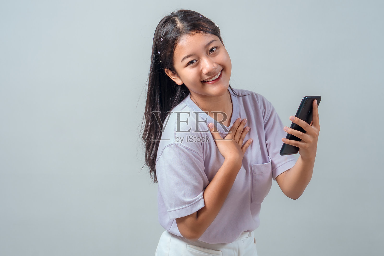 一位笑容灿烂的亚洲少女，身穿休闲服饰，在影棚内，一边开心地笑着，一边拿着手机，并用手触摸胸口，表达感激之情。照片摄影图片