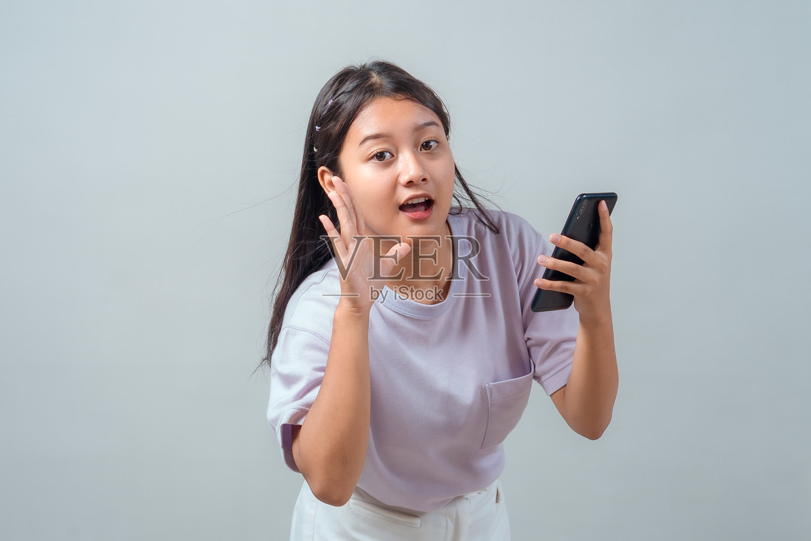 一位惊讶的年轻亚洲女性，她拿着智能手机，捂着耳朵，好像在仔细听。她穿着一件粉彩色T恤，表现出对声音的好奇或反应。照片摄影图片