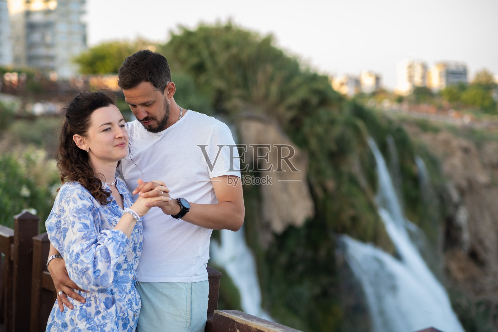 土耳其安塔利亚，情侣在日落时分，背景是城市天际线，在壮观的瀑布旁享受欢乐时光照片摄影图片