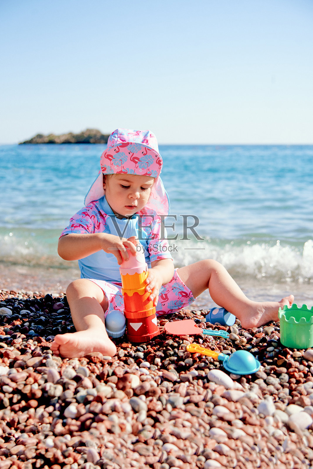 穿着蓝粉色泳衣的小女孩在海边鹅卵石沙滩上玩彩色玩具照片摄影图片