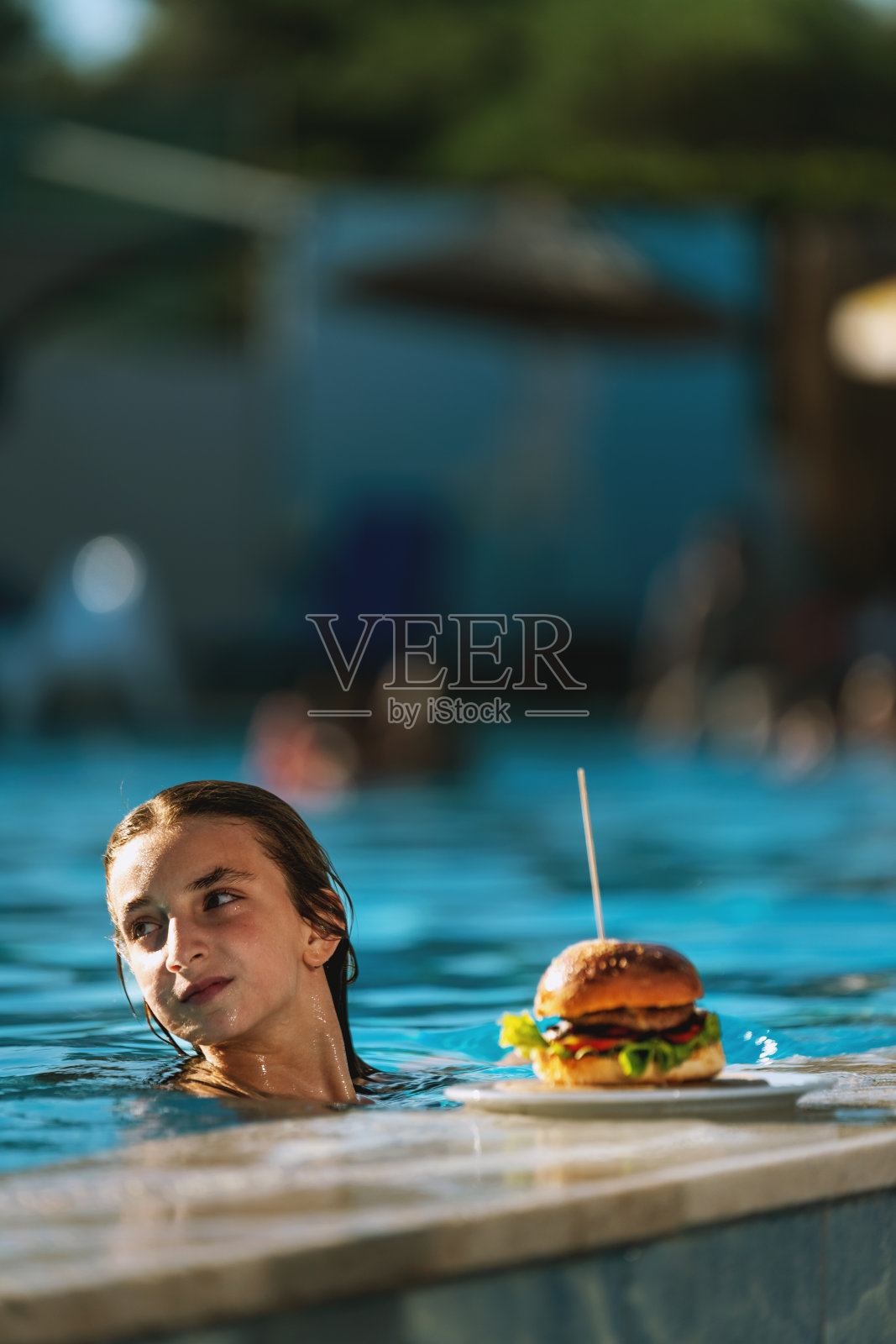 一个女孩坐在泳池边，开心地享用着多汁的汉堡照片摄影图片