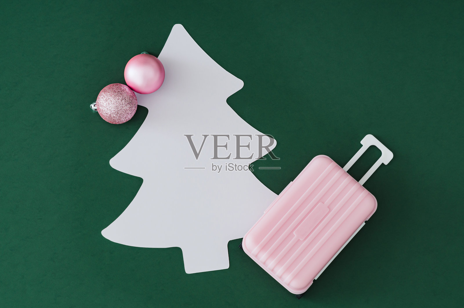 粉色行李箱、装饰球和圣诞树造型卡片在绿色背景上的圣诞场景，留有复制空间。简约时尚的旅行概念。潮流新年冒险创意。俯视拍摄。照片摄影图片