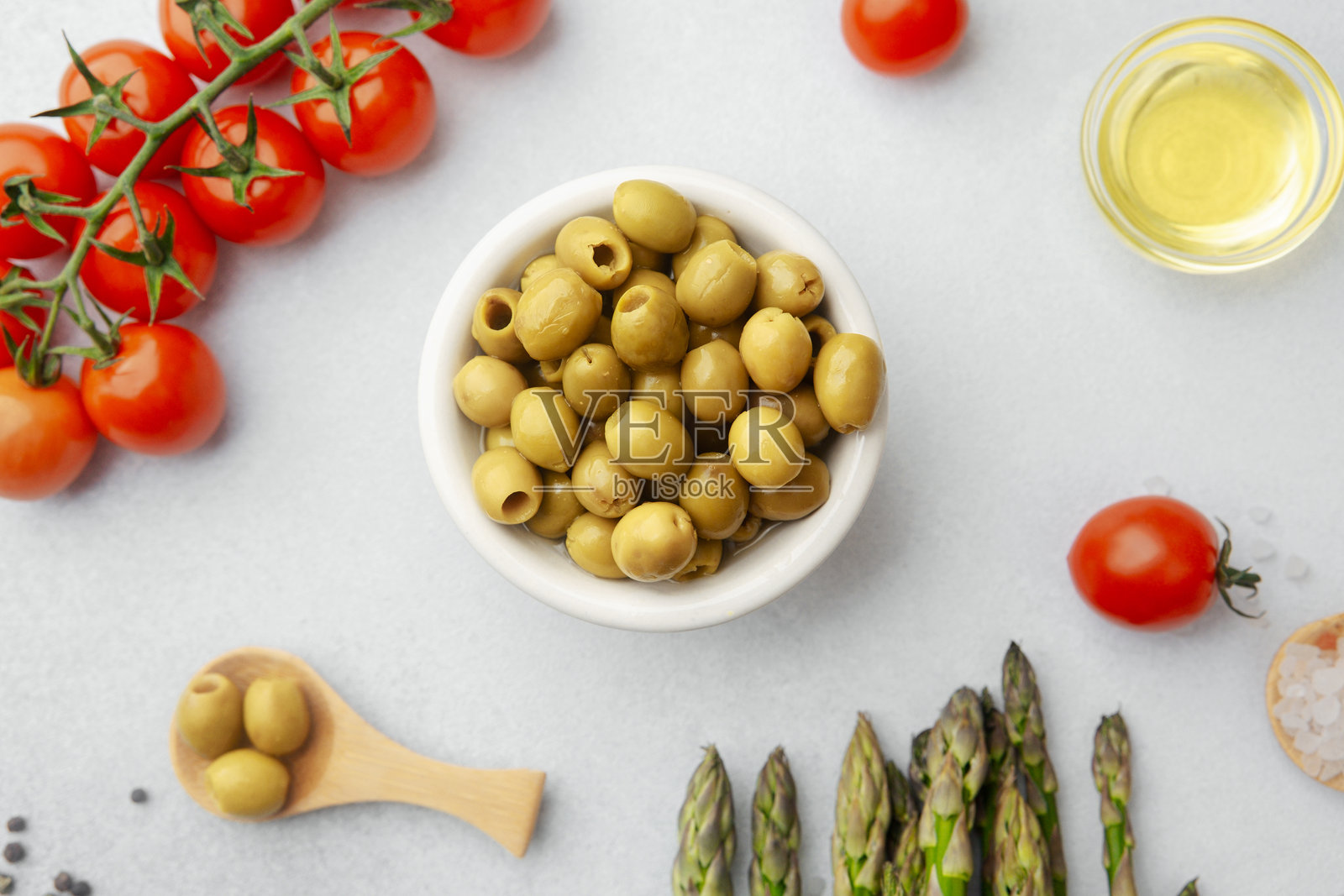 白桌上，陶瓷碗里盛着绿橄榄，旁边还有番茄、芦笋和调味料。照片摄影图片