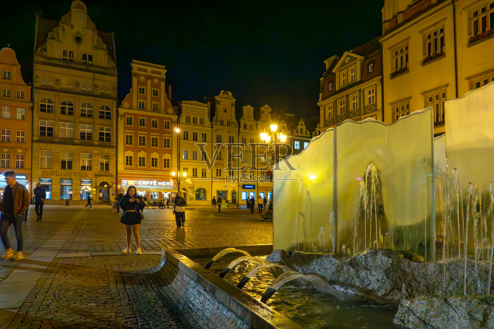 弗罗茨瓦夫——市场广场上的玻璃喷泉在夜晚，周围有联排别墅和行人。照片摄影图片