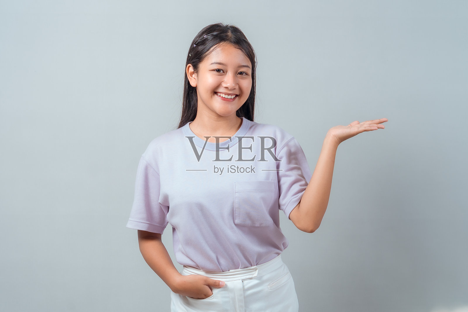 一位年轻的亚洲女性，身着柔和色彩的服装，一手插在口袋里，另一手掌心向上，随意地站着，仿佛在展示或呈现某物，背景为灰色，人物被独立出来。照片摄影图片