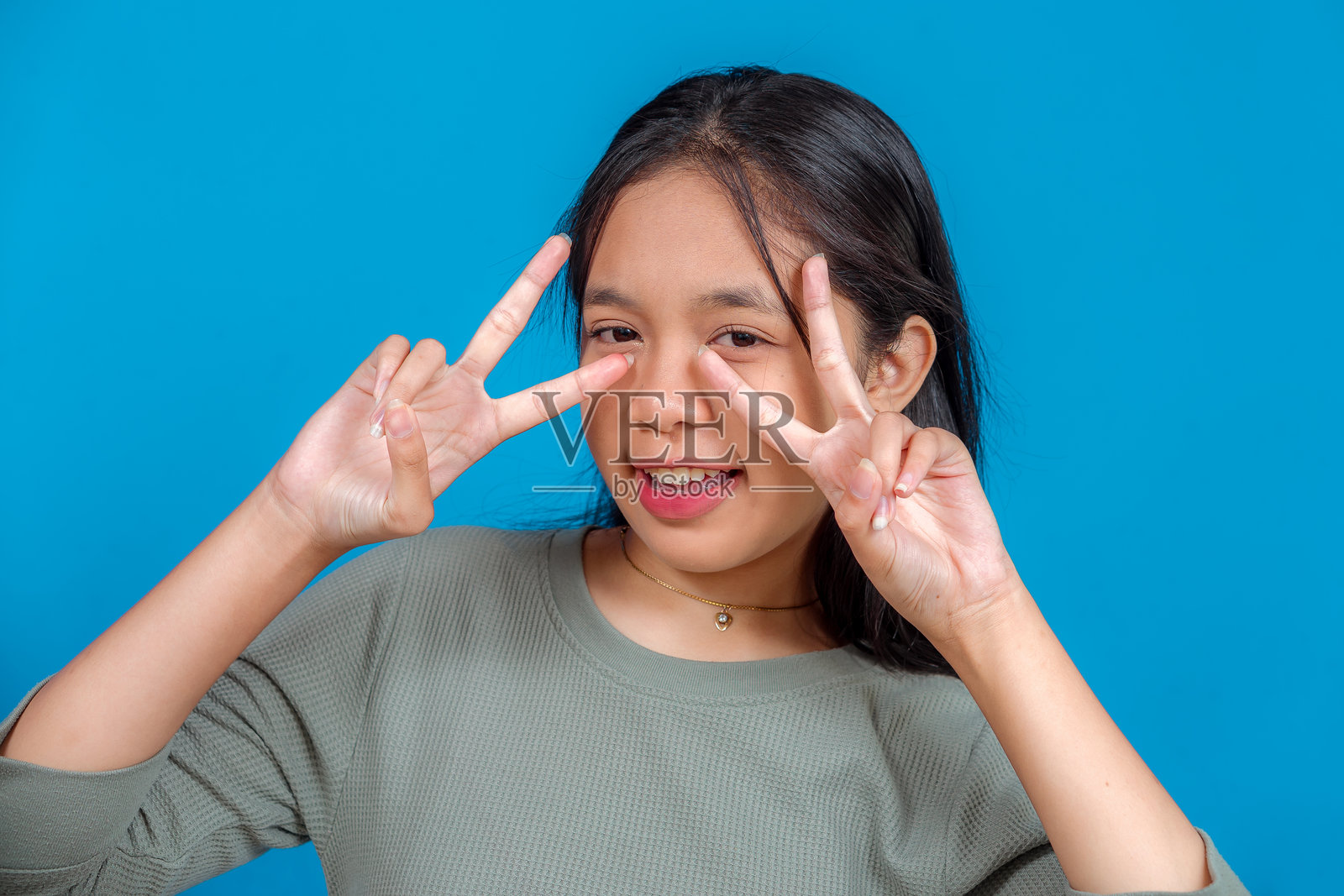 自信喜悦的年轻亚洲女孩，对着镜头俏皮地竖起双V手势，背景为蓝色，适合青年生活方式推广。工作室人像。照片摄影图片