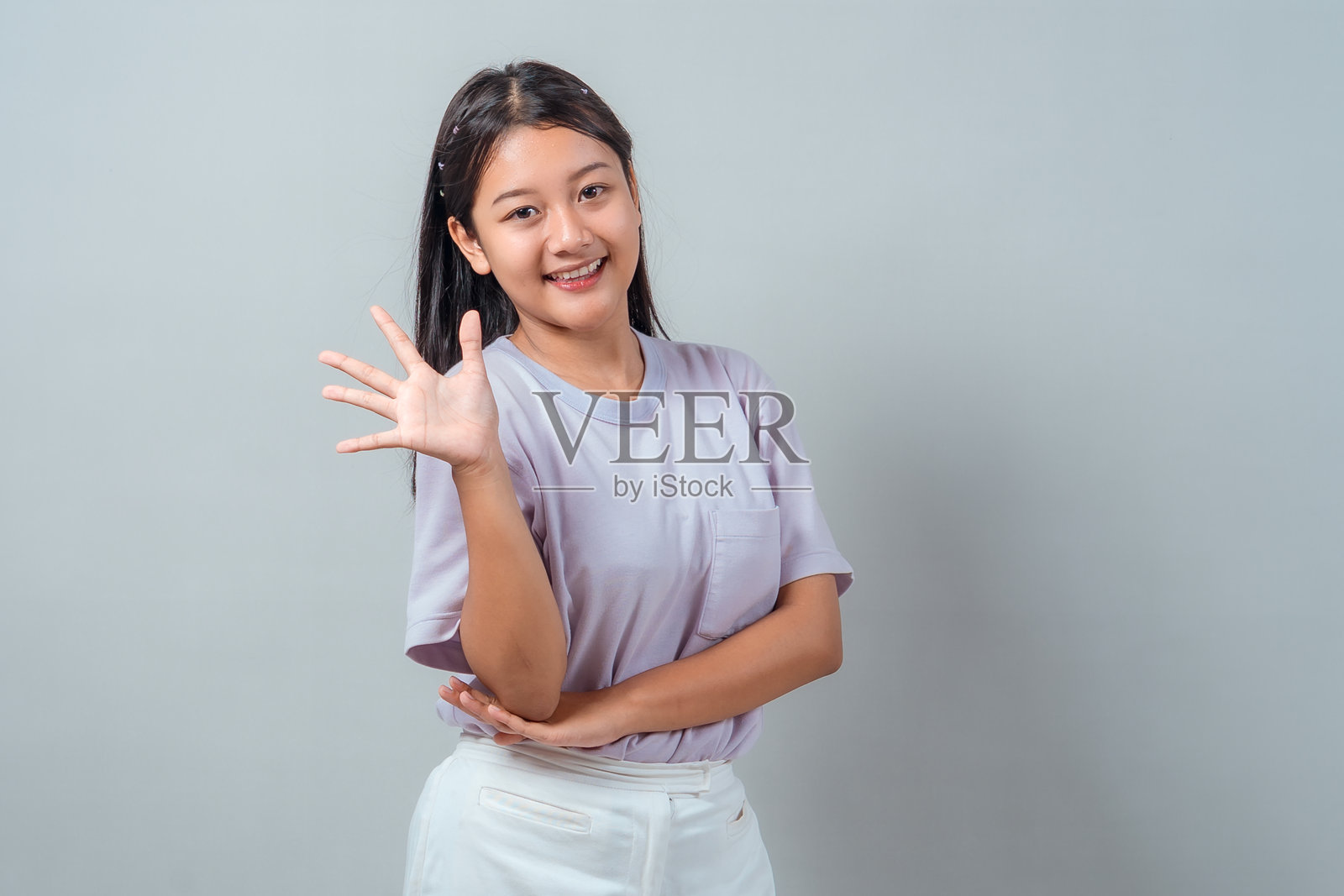 身穿浅紫色T恤的年轻亚洲女性肖像，面带微笑，张开手掌挥手致意，自信地站在纯灰色背景前。照片摄影图片