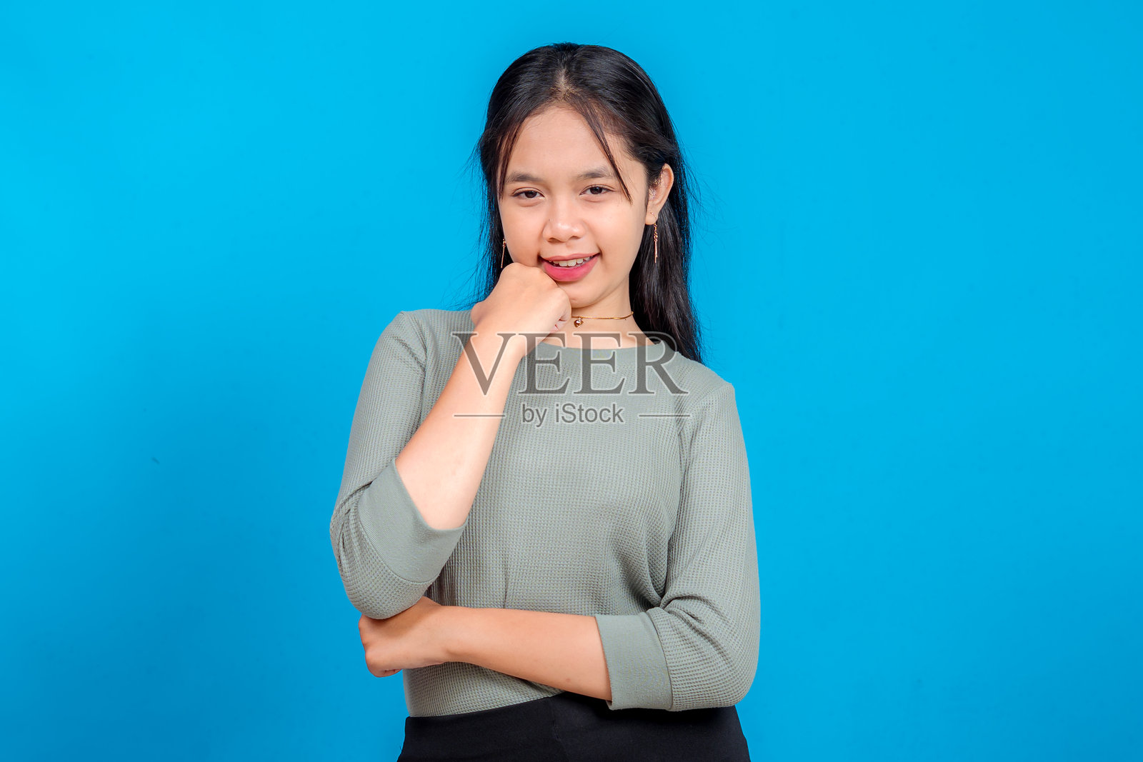 年轻的亚洲女性自信地微笑，表情俏皮，一只手托着下巴，双臂交叉，站在充满活力的蓝色背景前，直视镜头。照片摄影图片