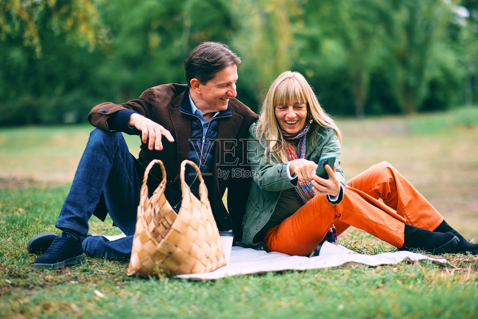 成熟的夫妇在公园享受秋季野餐，同时使用手机照片摄影图片