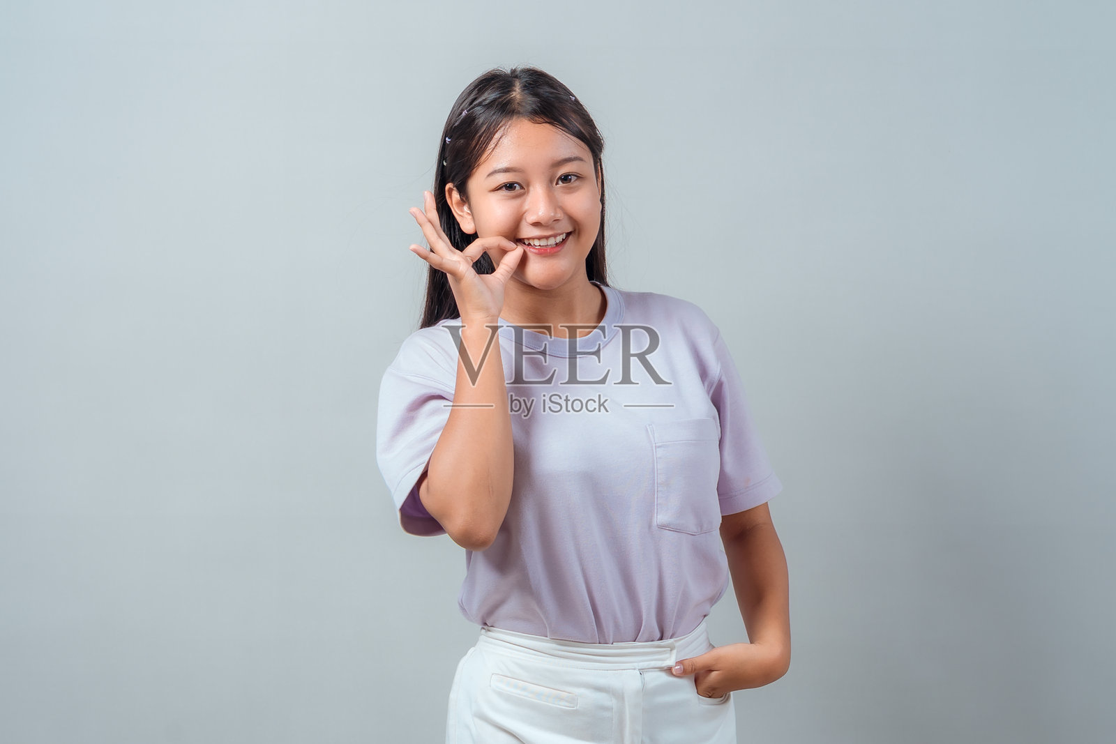 一个穿着淡紫色T恤和白色裤子的快乐亚洲小女孩，脸上带着灿烂的笑容，竖起了OK手势。自信地站立在纯净的浅色背景前。照片摄影图片