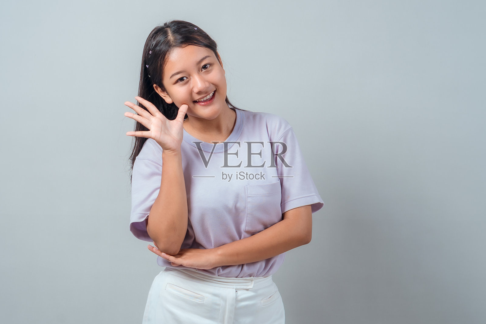 一位年轻的亚洲女性，身穿淡紫色T恤，面带微笑，友好地挥手致意，她休闲地站立在纯灰色背景前，代表着问候或你好。照片摄影图片