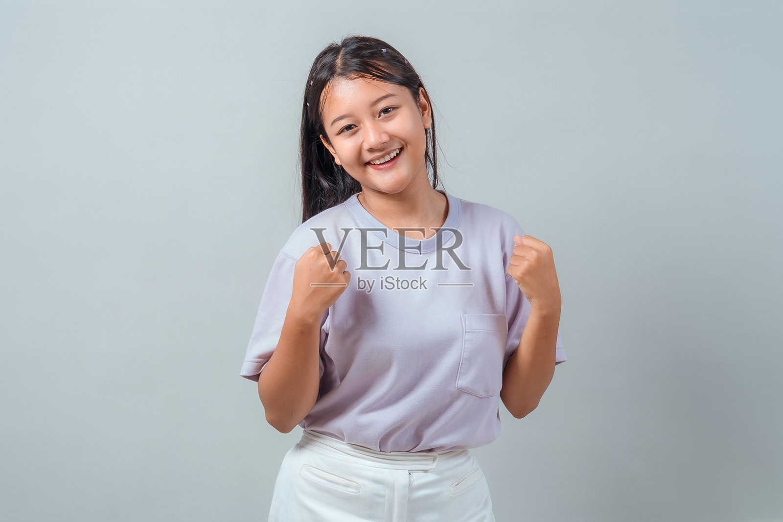 一位快乐的年轻亚洲女性，面带微笑，自信地举起双拳。她身穿淡紫色T恤和白色裤子，站在中性的灰色背景前。照片摄影图片