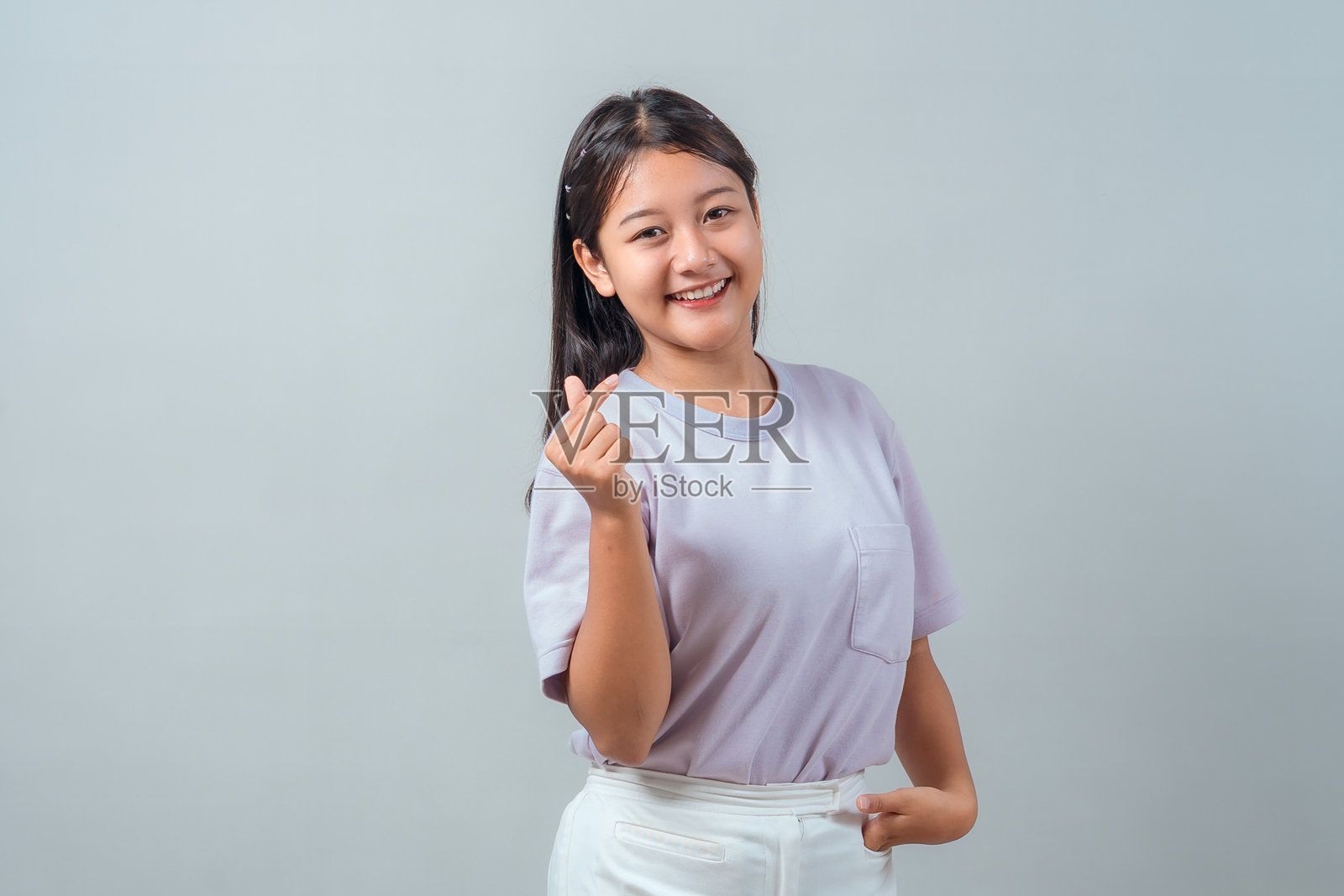 一位穿着浅紫色T恤和白色裤子的快乐亚洲年轻女性，面带幸福的微笑，在工作室的纯色背景前，比出韩式手指比心手势。照片摄影图片