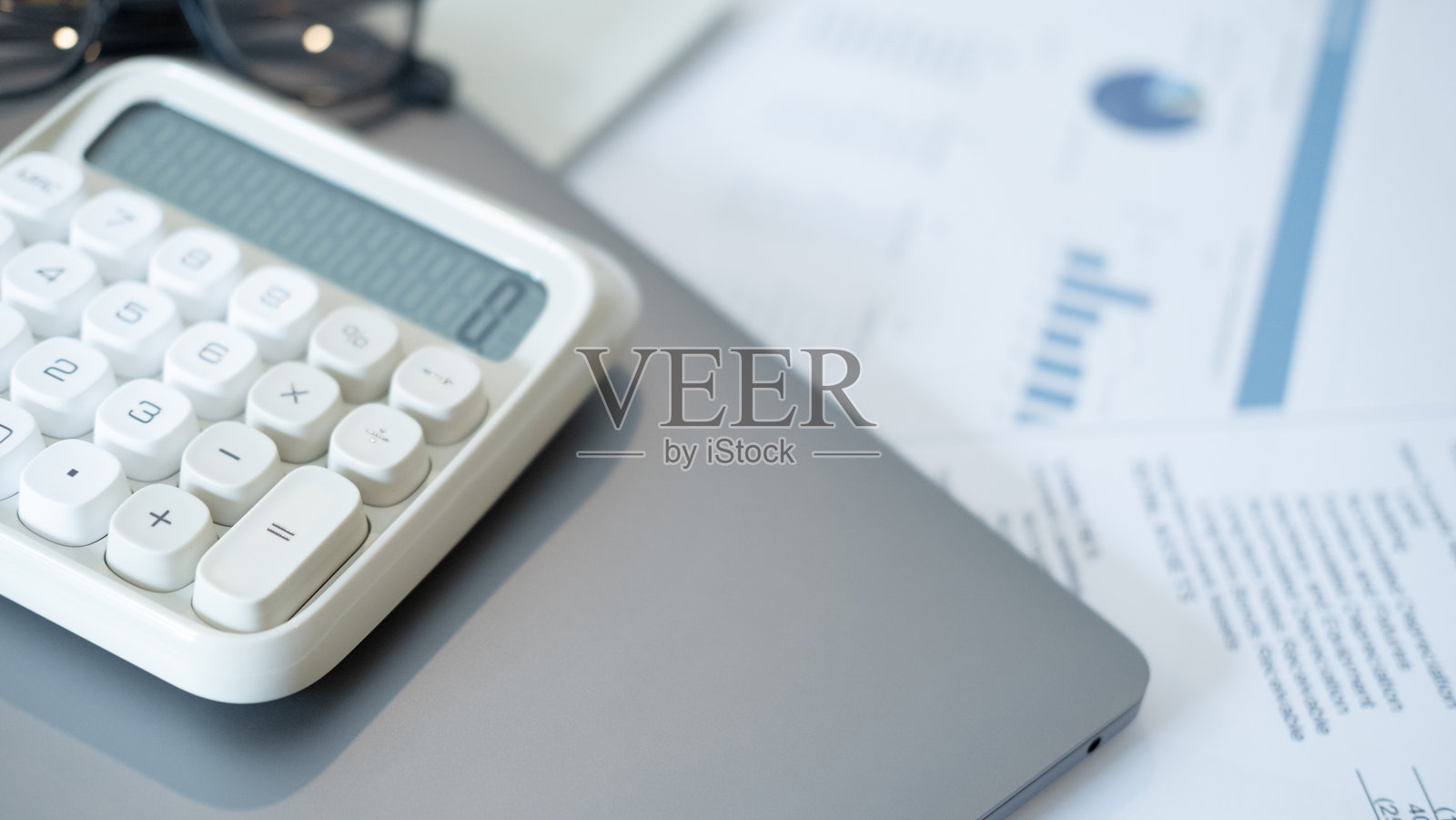白色的计算器，带有大按钮，放在灰色笔记本电脑旁，旁边是带有蓝色图表的财务文件，放在木制桌子上，象征着财务规划和分析。照片摄影图片