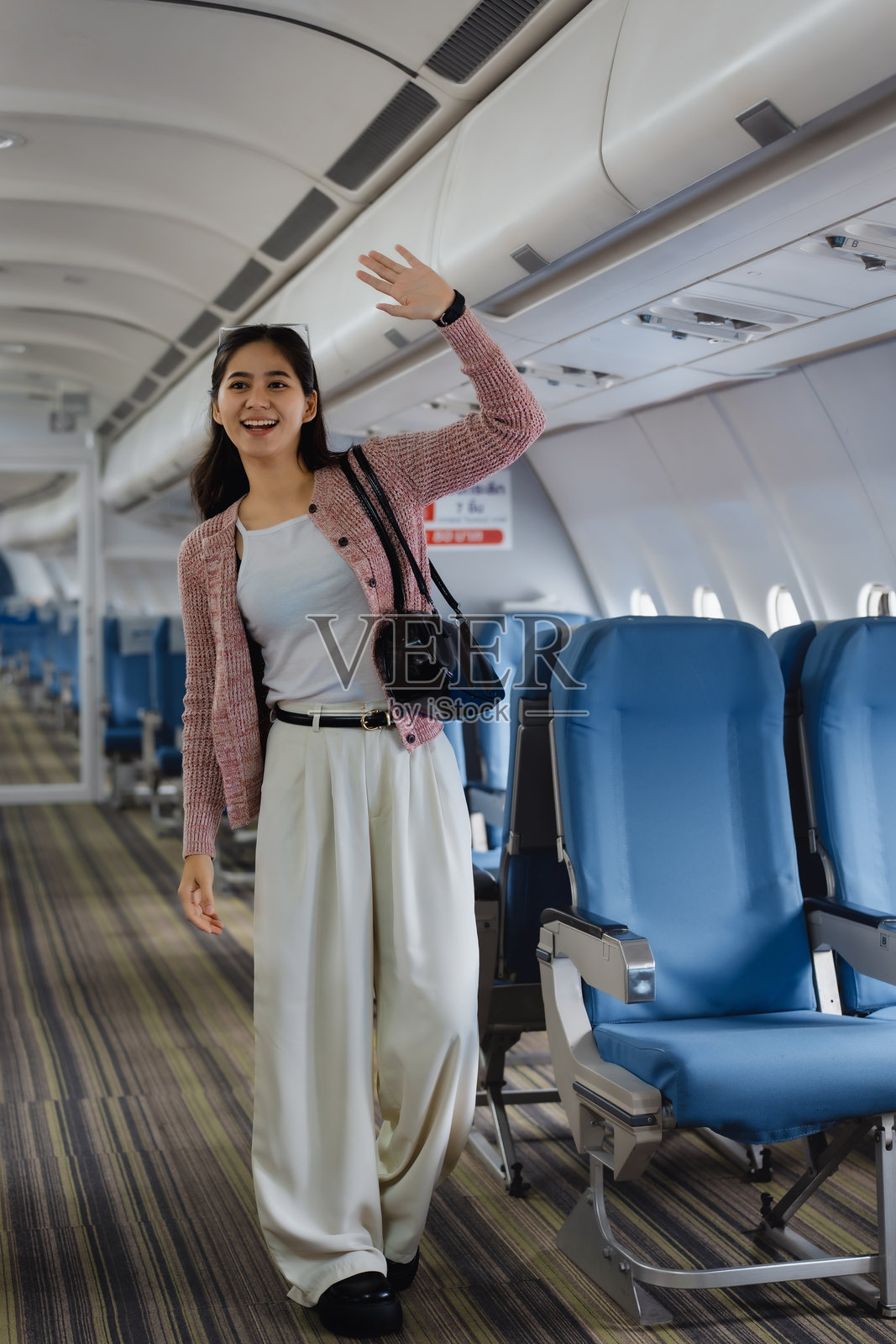 一位亚洲女性坐在飞机窗边，穿着休闲粉色毛衣和白色裤子，脸上洋溢着温暖的笑容，在舒适的机舱旅途中散发出轻松自信的气息。照片摄影图片
