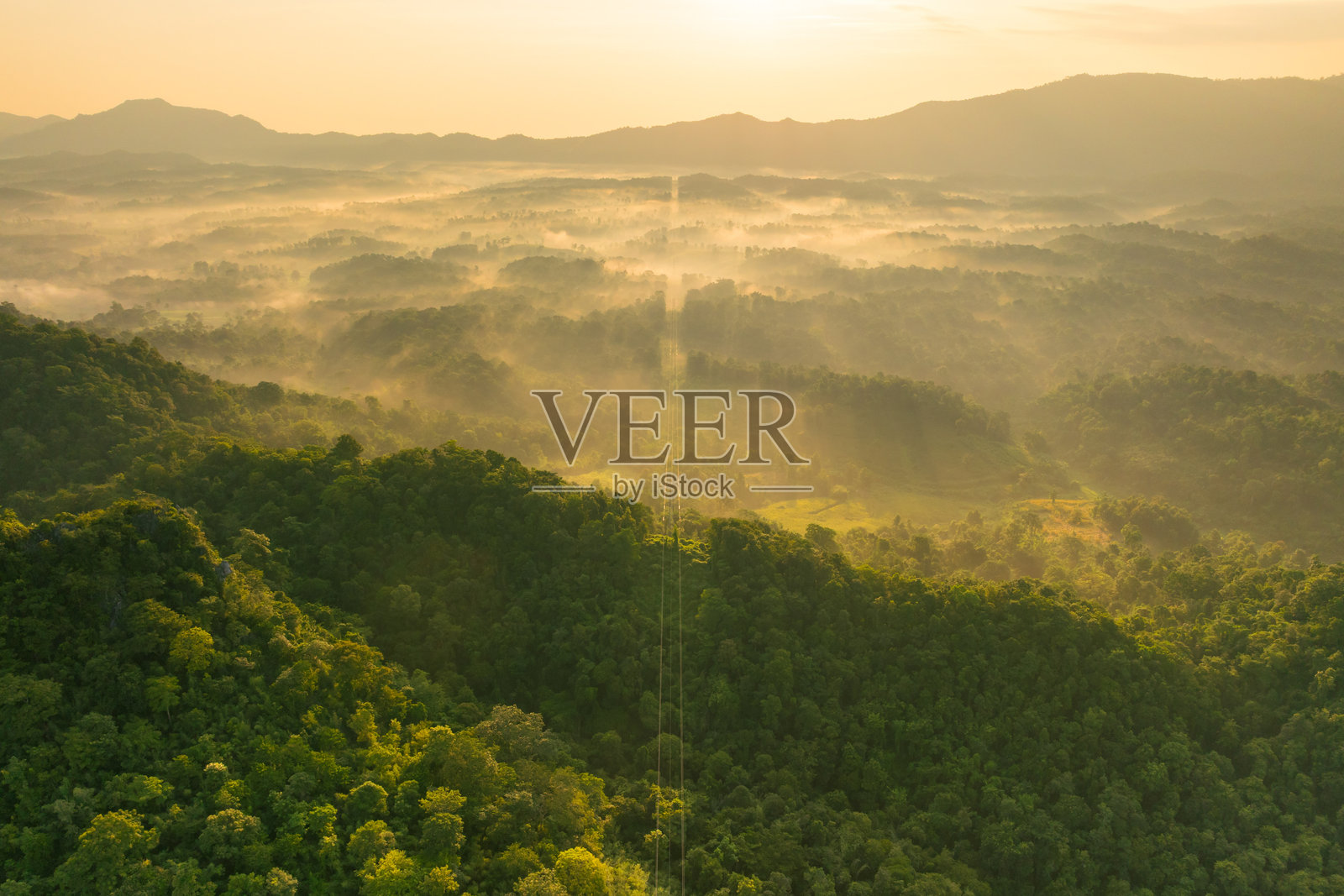 日出时分，无人机航拍绿色山峦上的电力线和输电塔。晨雾营造出令人惊叹的背景，金色的太阳为基础设施投下美丽的光辉。照片摄影图片