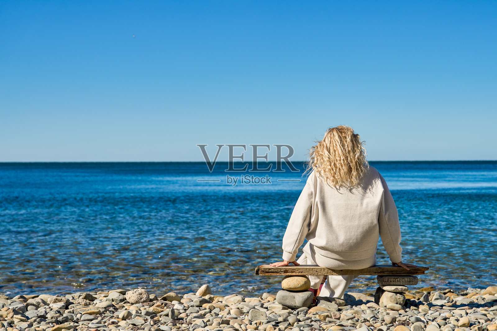 一位金发成熟的女性的背影，她正悠闲地坐在海边，欣赏宁静的氛围，享受放松的时刻，留有复制空间，概念为户外休闲和海洋生态系统。照片摄影图片