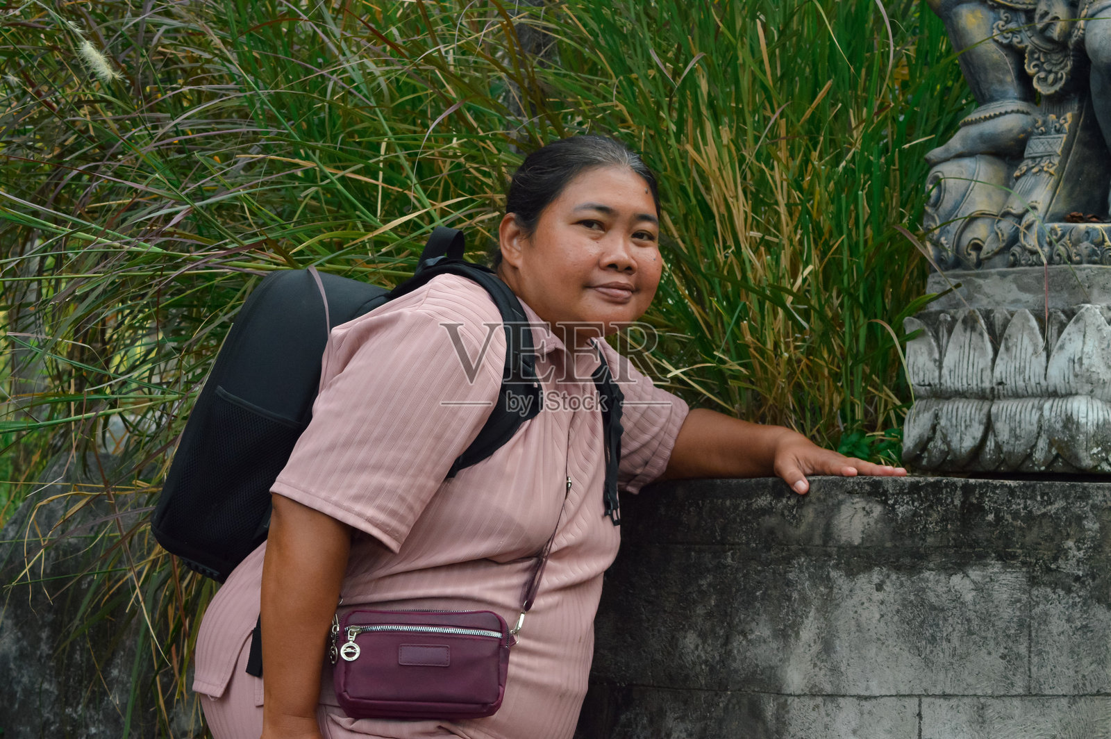 身穿白色长裙的女子倚靠在古老的石头墙边，旁边有一尊小型雕像。照片摄影图片