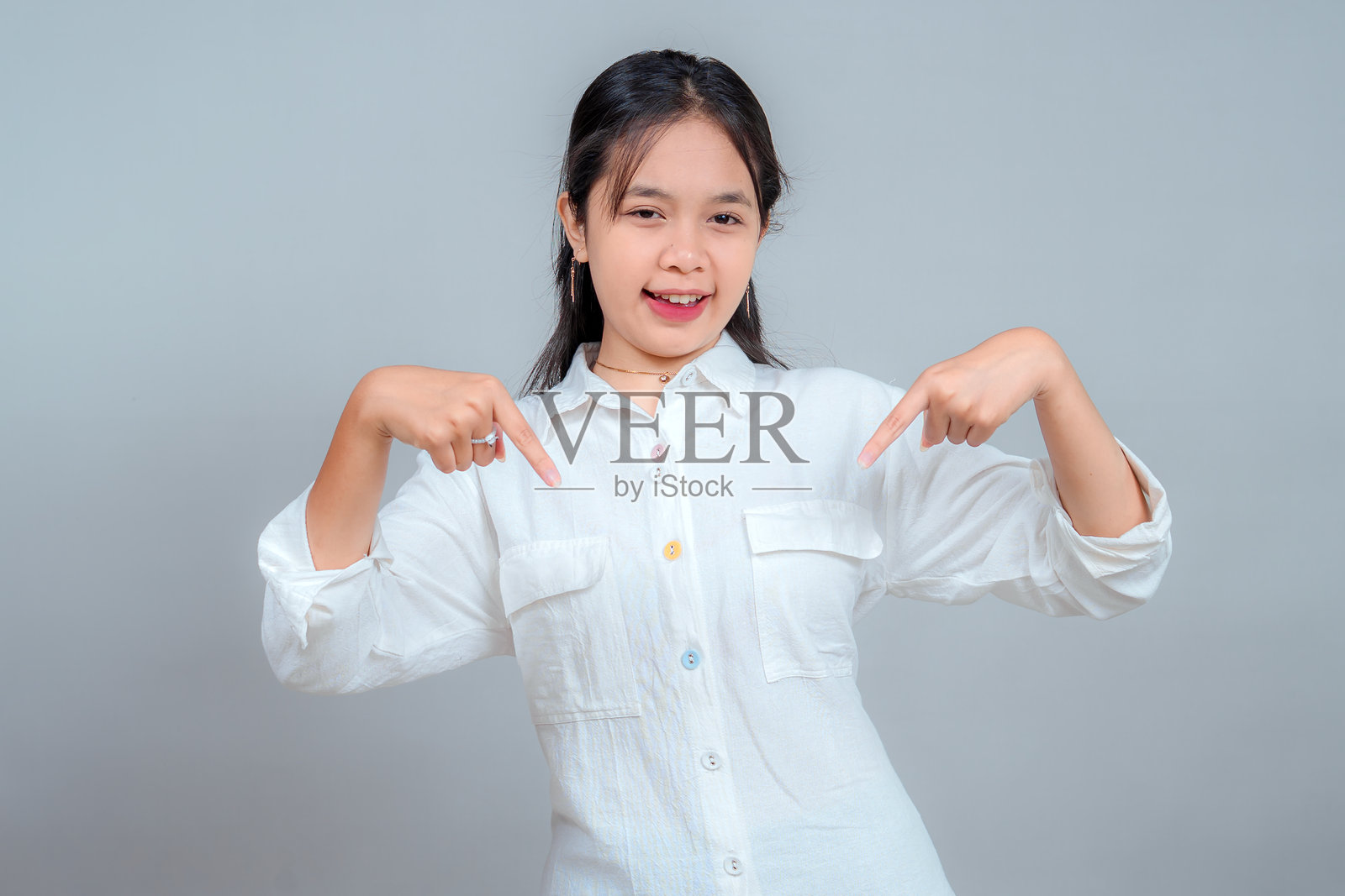 自信的亚洲女孩穿着白衬衫，面带微笑，双手食指指向下方，表达兴奋并吸引注意，孤立在灰色背景上供宣传使用。照片摄影图片