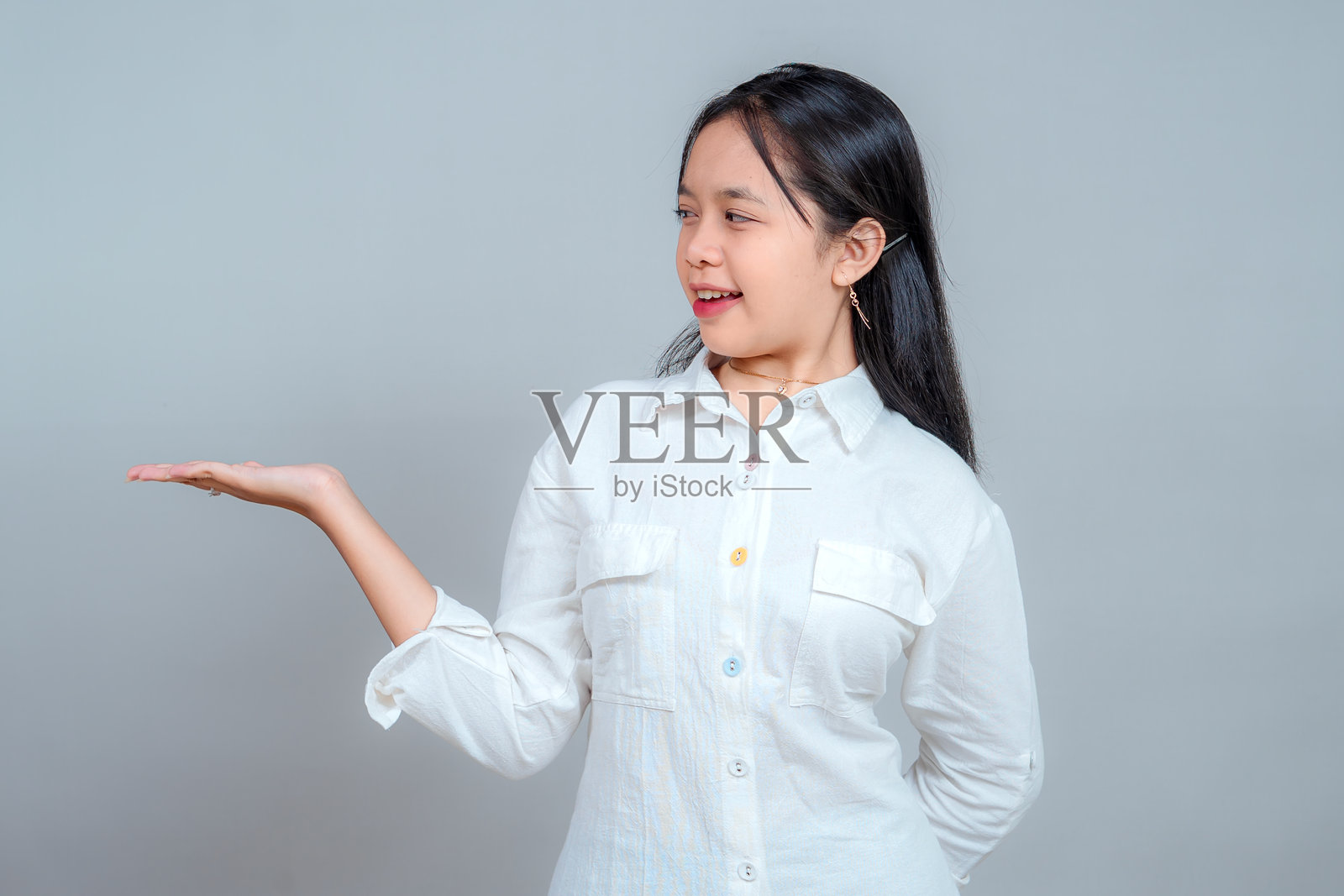 快乐的亚洲女孩，穿着白衬衫，微笑着，张开手掌指向一侧，带着兴奋的表情看着自己的手，暗示产品放置，孤立在灰色背景上。照片摄影图片
