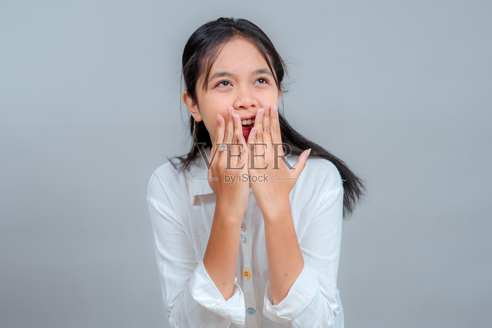 一位表情惊讶、张着嘴、双手靠近嘴唇的年轻亚洲女性。工作室拍摄，灰色背景，充满喜悦的情感反应。人类情感概念。照片摄影图片