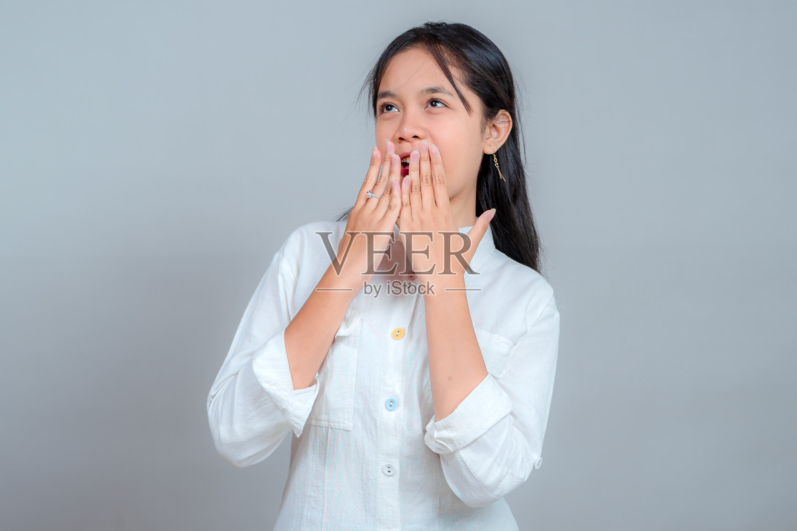 年轻亚洲女性的肖像，她抬头惊叹又欣喜地张着嘴，双手捂着嘴唇。在灰色背景下的影棚拍摄。情感反应概念。照片摄影图片