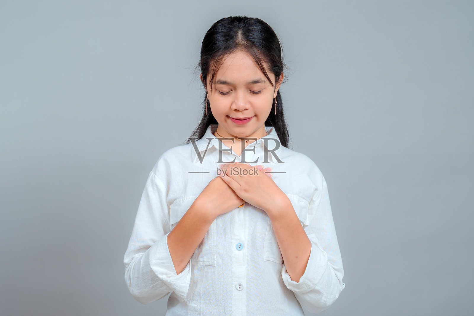 一位亚洲少女微笑着闭上眼睛，双手放在胸前，表达出平静、真诚和感激之情。非常适合描绘情感丰富、发自内心的时刻。照片摄影图片