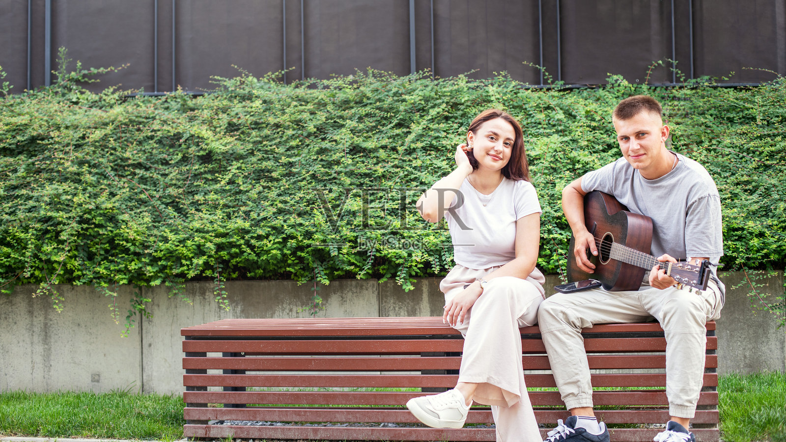 一对快乐的年轻情侣坐在公园长椅上，一起弹吉他，男人教女人弹奏，享受音乐，共度户外时光。照片摄影图片