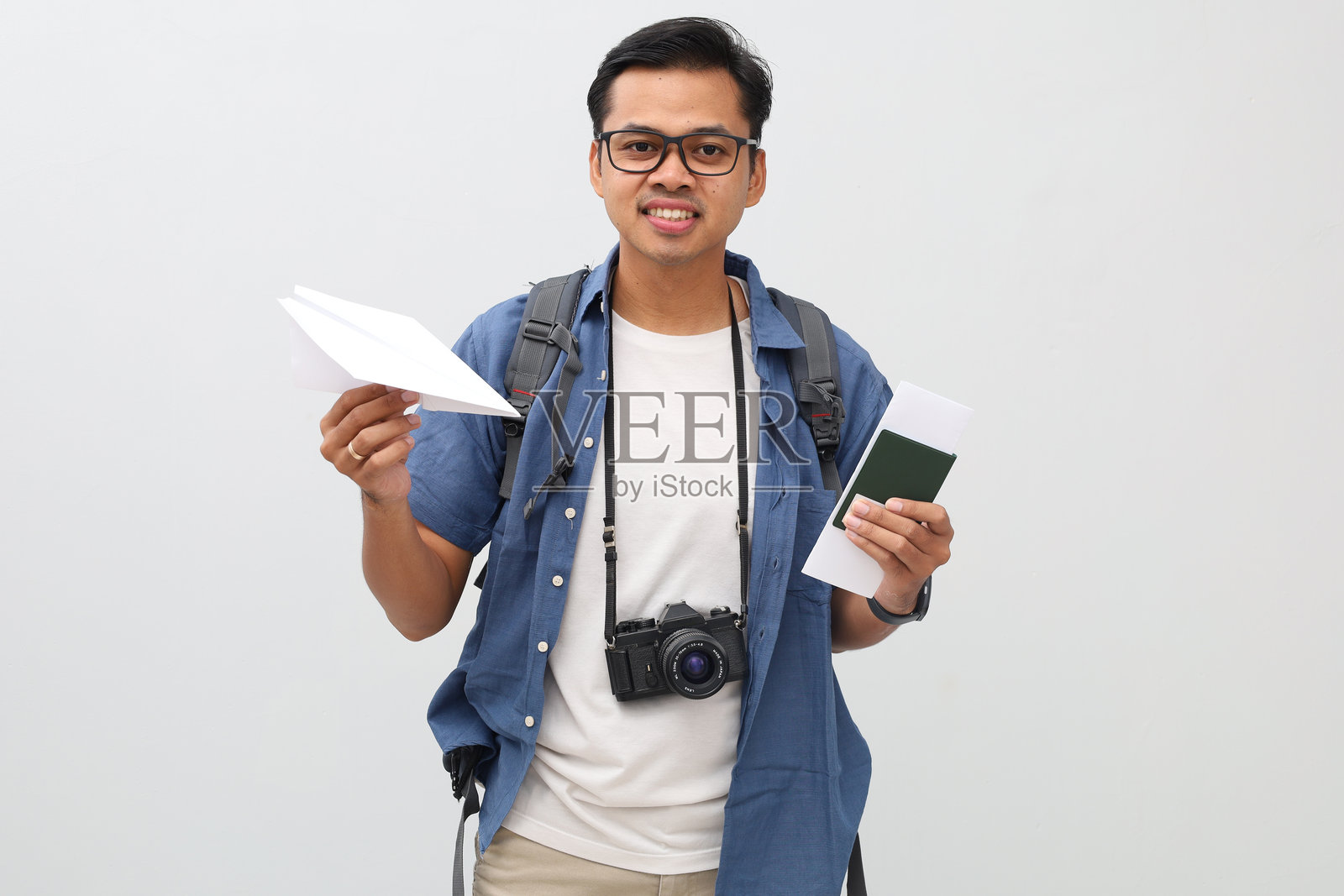 一位男游客，身着休闲装，背着背包，脖子上挂着相机，手持护照和机票以及飞机模型，体现航空旅行的概念，他站着，微笑着看向镜头。照片摄影图片