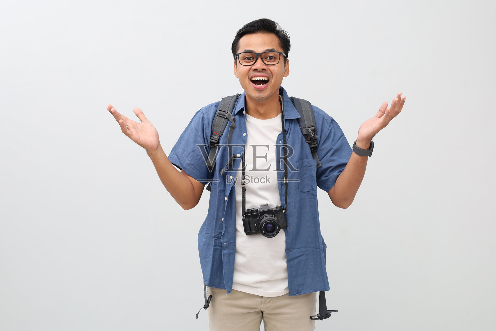 一个年轻的亚洲男子，穿着休闲服，背着背包，脖子上挂着相机，惊讶地看着某物，双手高举，快乐地大喊，孤立在白色背景上，度假概念。照片摄影图片