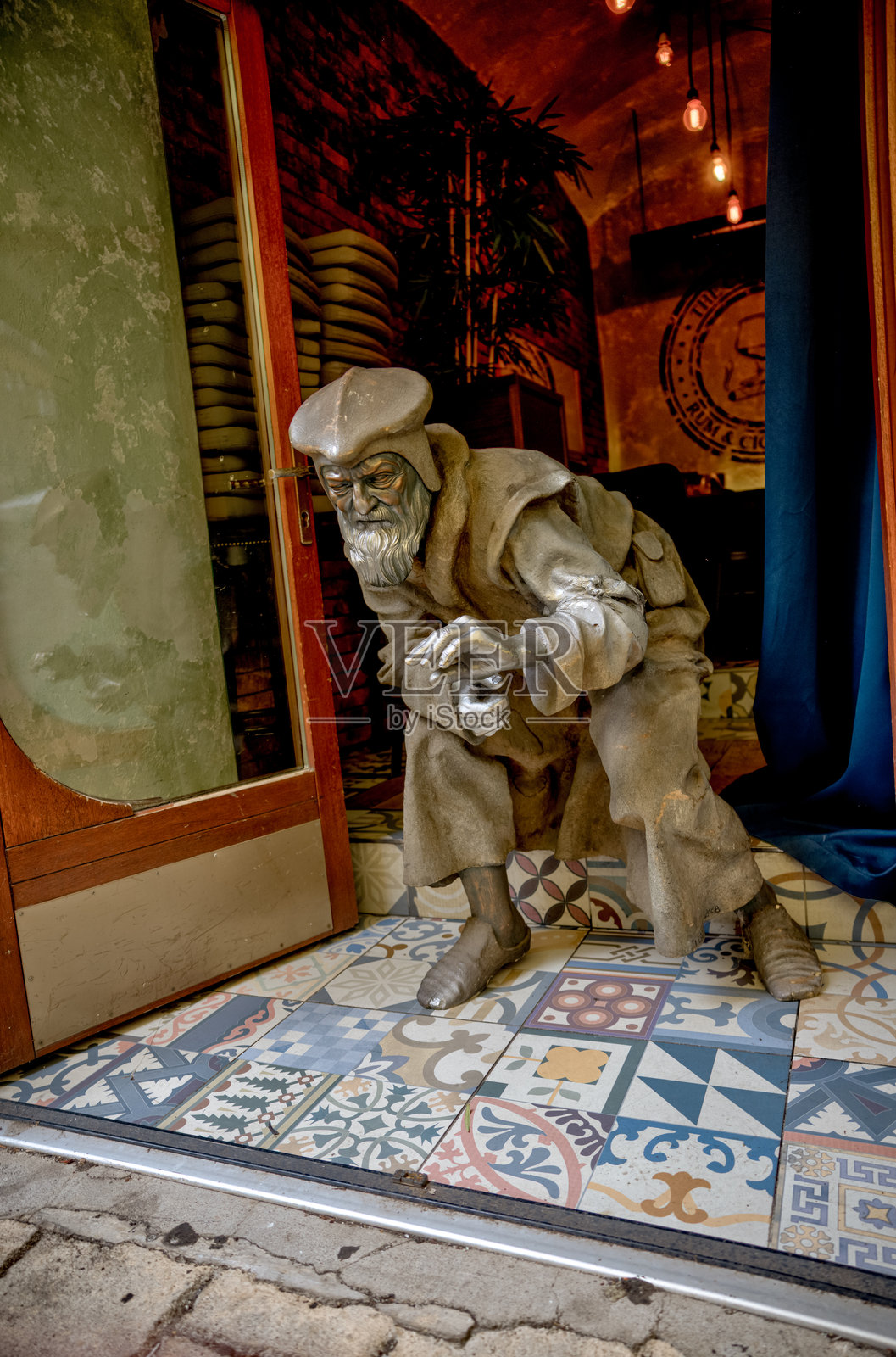 一个真人大小的驼背中世纪人物雕塑在当地一家商店的入口处迎接访客，它矗立在色彩鲜艳的图案瓷砖上。艺术家和创作日期不详。照片摄影图片