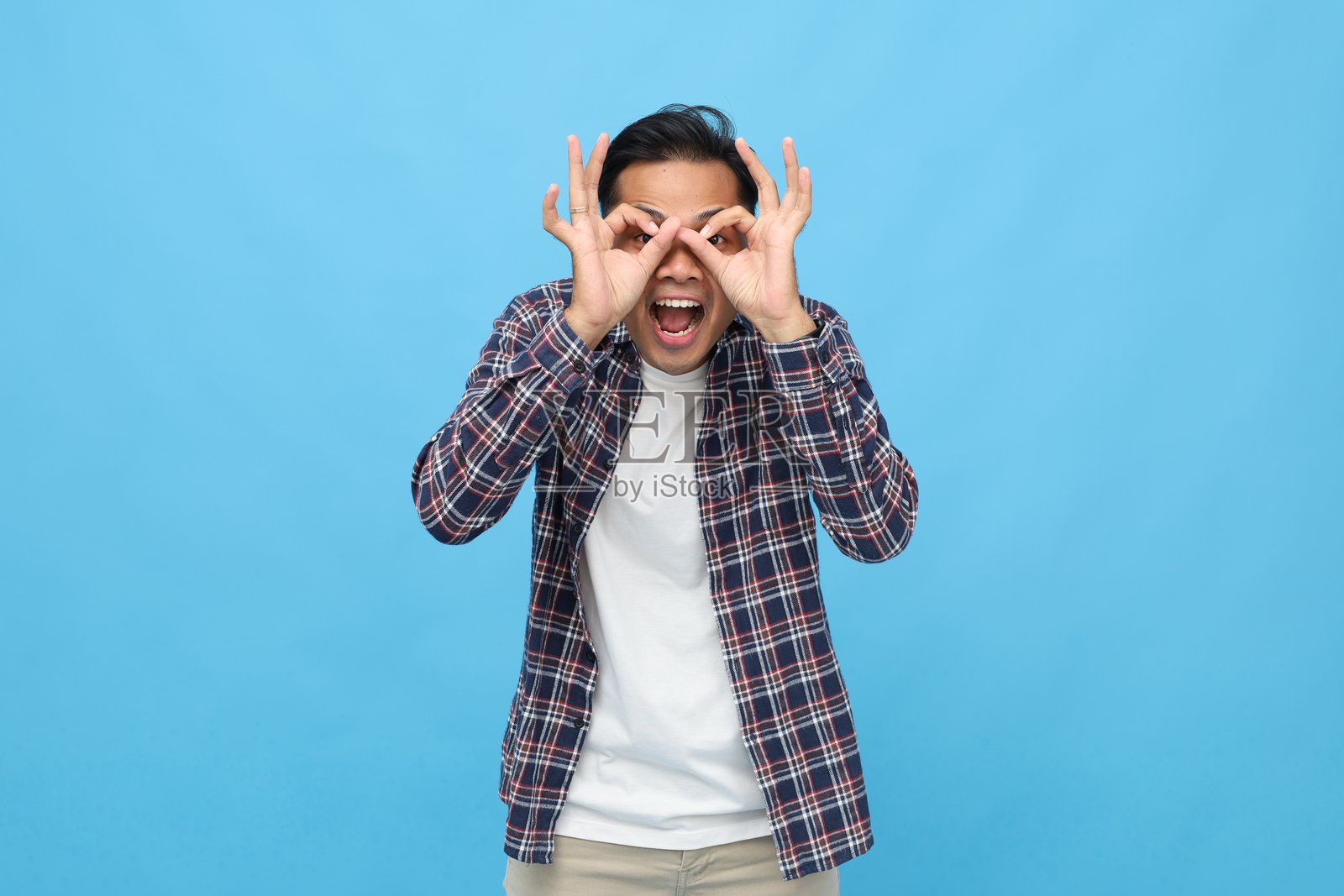 年轻亚洲男子穿着休闲服，手势比作望远镜，对着镜头大喊，背景为蓝色。照片摄影图片