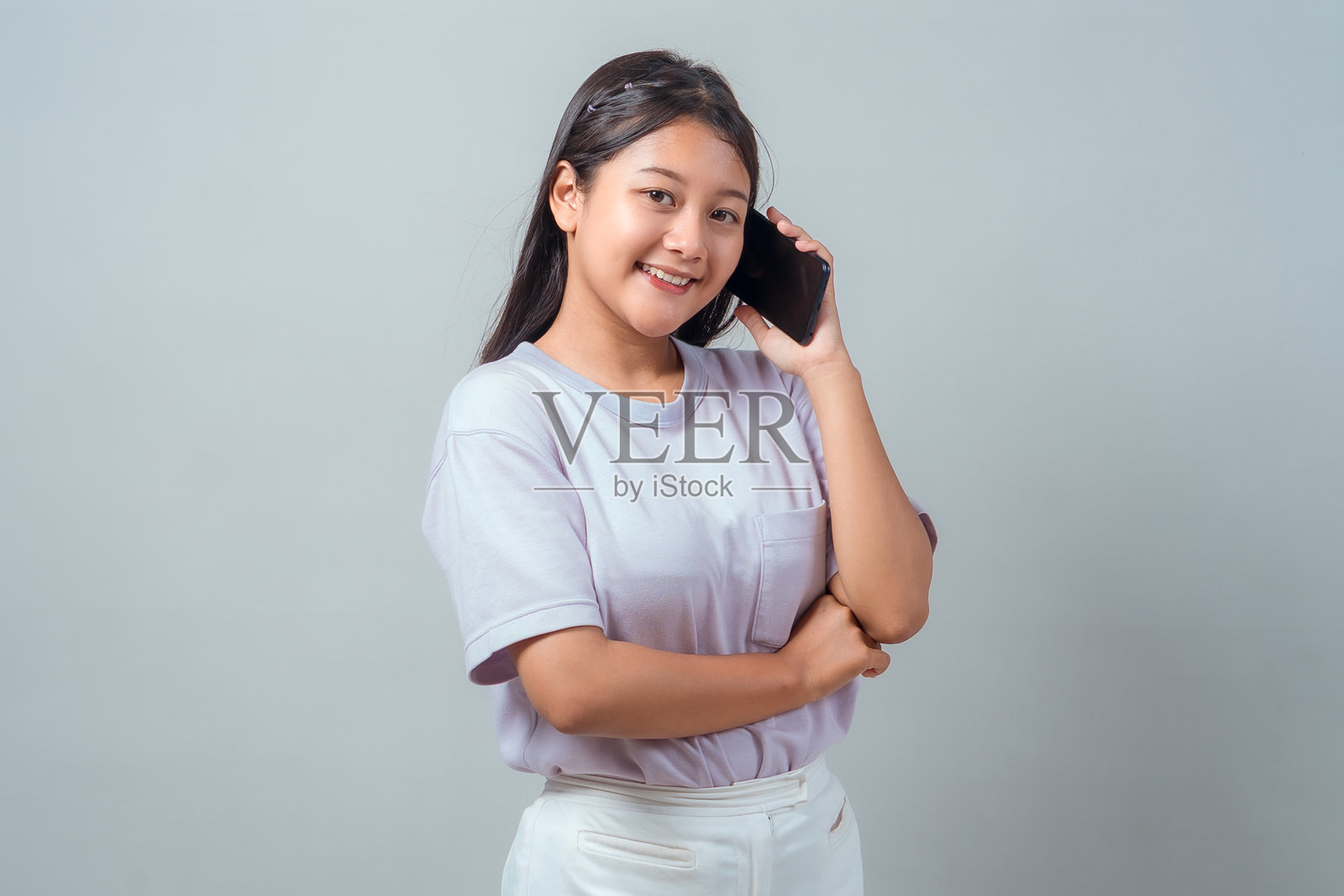 自信的年轻亚洲女性，身穿休闲服饰，单臂交叉站立，面带微笑地通过手机交谈。纯色背景下的工作室肖像，用于沟通概念。照片摄影图片