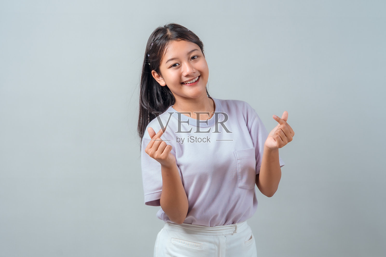 快乐的年轻亚洲女性微笑着做出双重韩式手指心形手势。她穿着淡紫色T恤和白色裤子，在纯色背景的影棚里开心地摆姿势。照片摄影图片