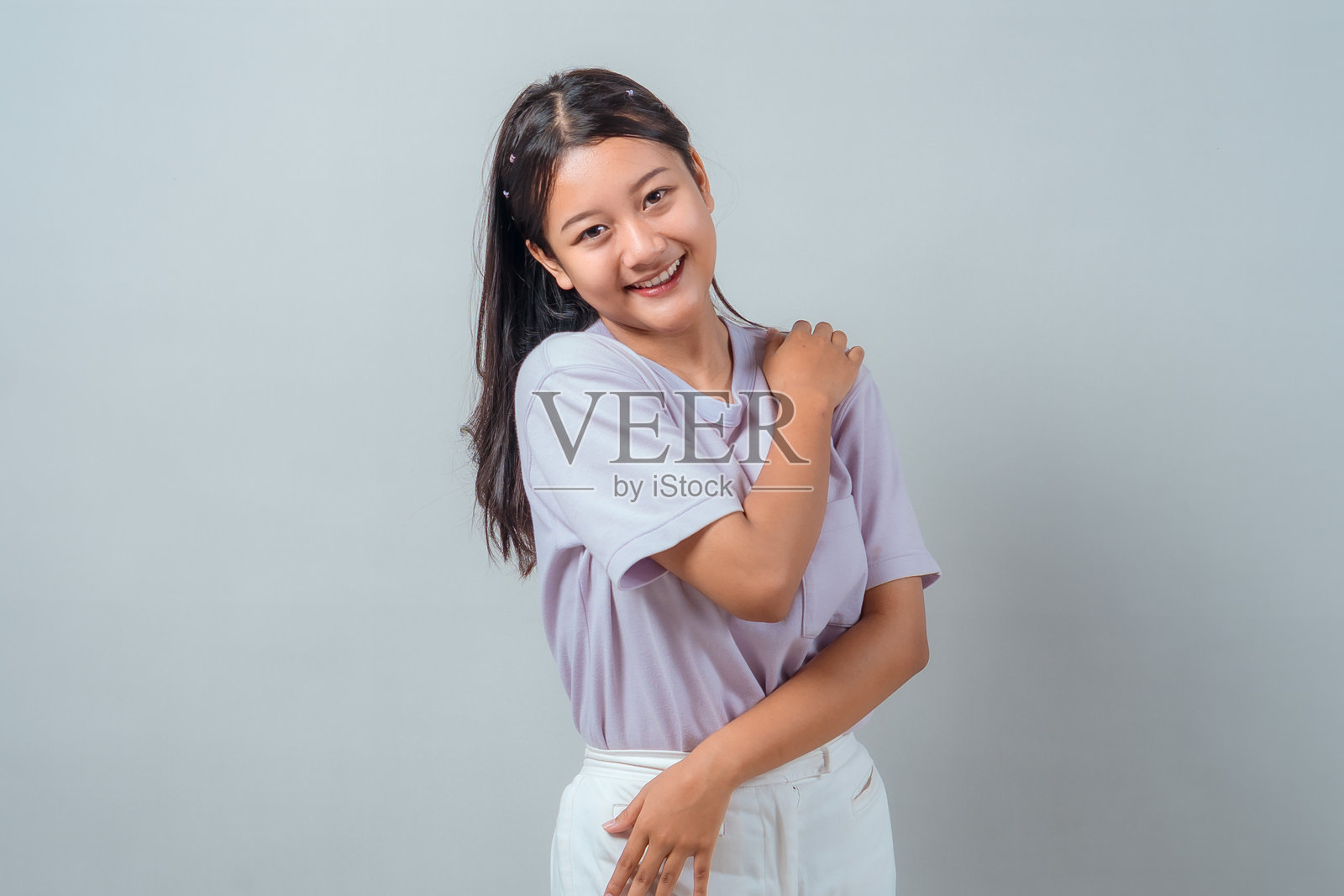 一位年轻的亚洲女性面带微笑，姿势放松，轻轻触碰自己的肩膀，身穿休闲的淡紫色衬衫和白色裤子，站在纯色背景前。照片摄影图片