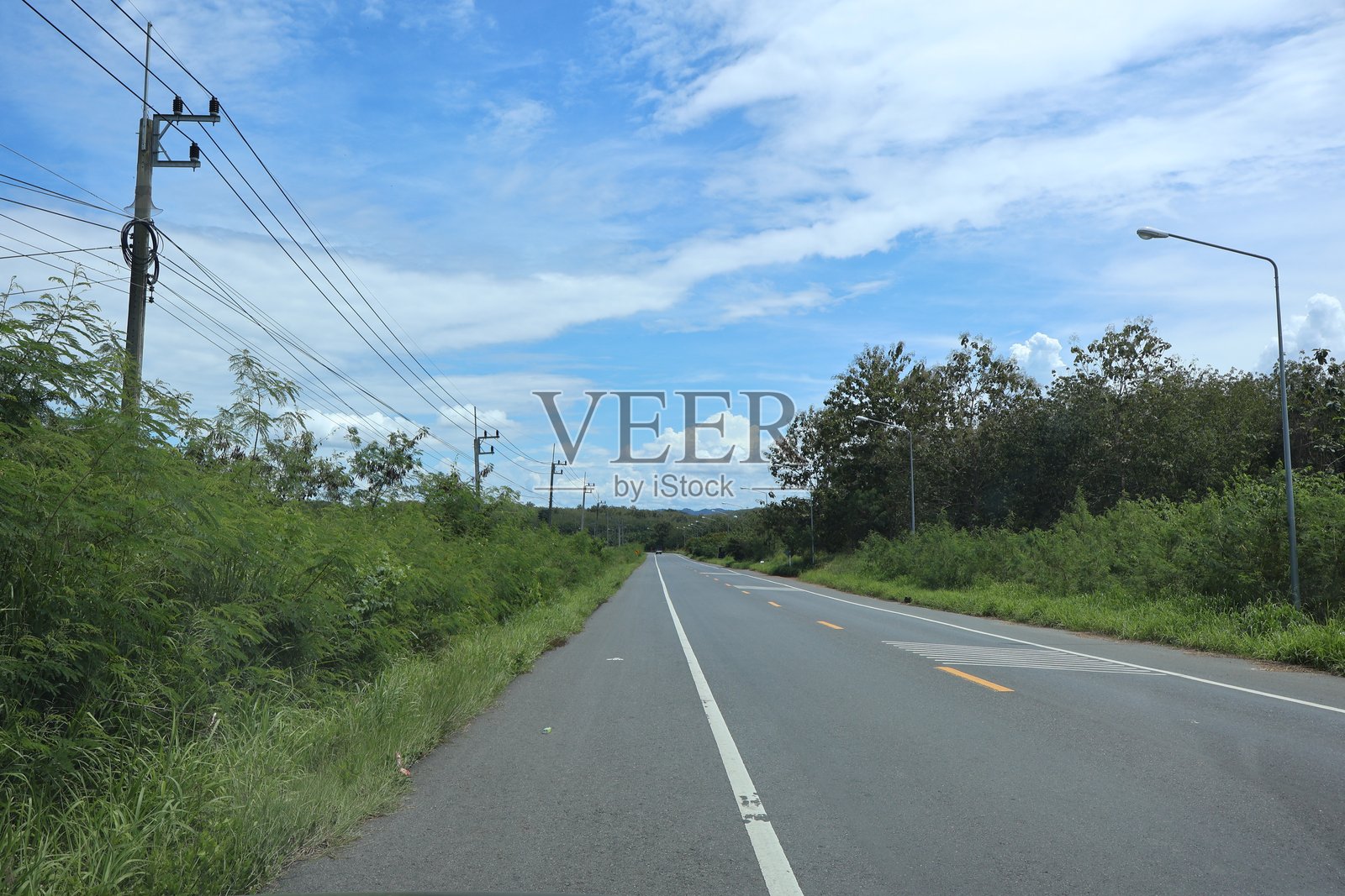 阴天下的乡村道路向前延伸，路的一侧是茂密的绿植，另一侧的栅栏与贯穿画面顶部的电线平行。照片摄影图片