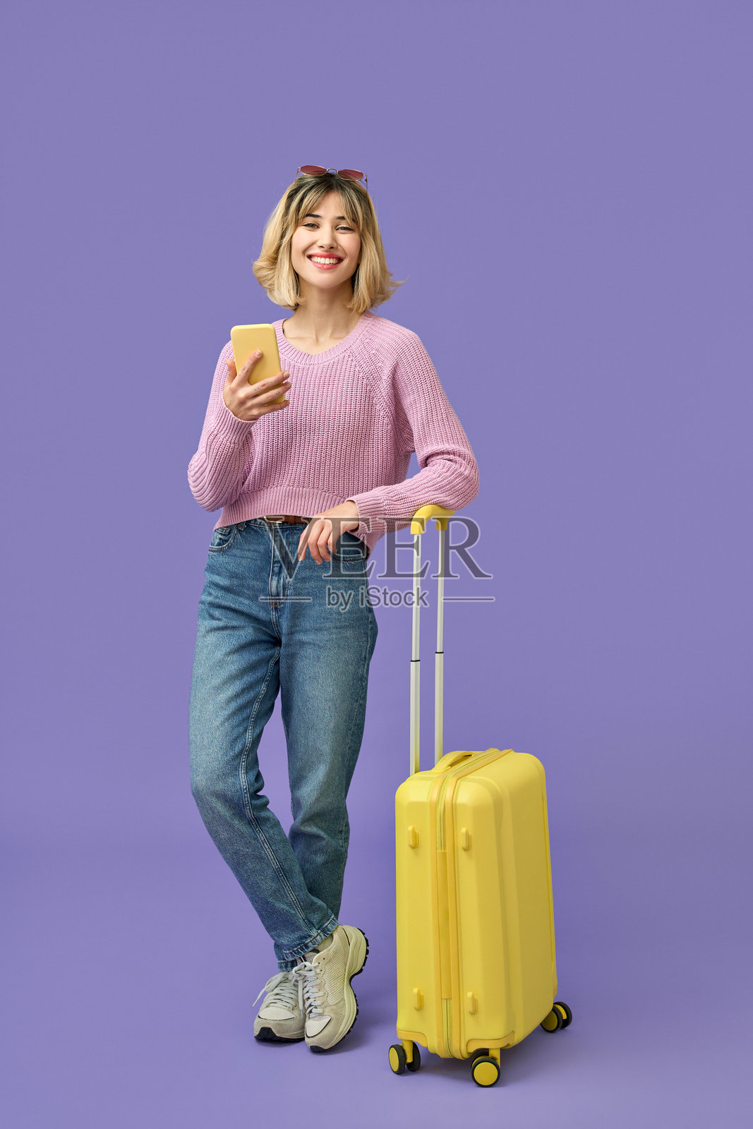开心的女孩拎着行李箱，一边使用手机，独自站在紫色背景前。照片摄影图片