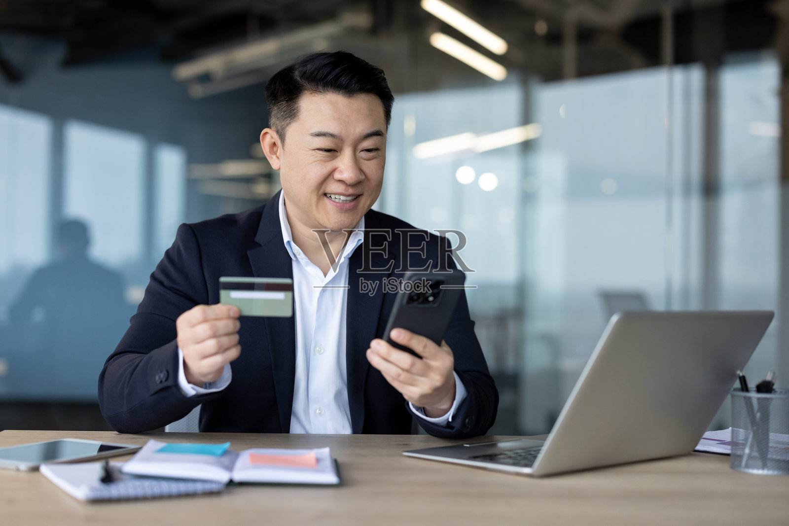 一位年轻的亚洲男子微笑着坐在办公室的办公桌前，使用手机和信用卡。照片摄影图片