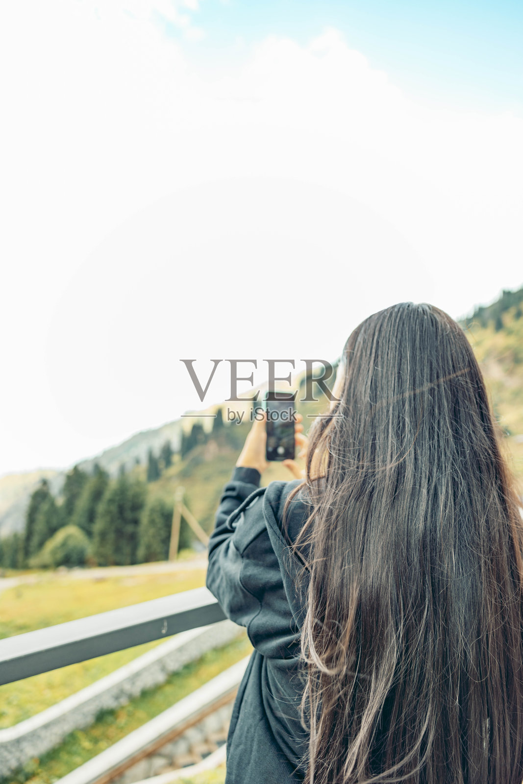 一位留着黑色长发的年轻亚洲女性正在拍摄山景。画面展示了郁郁葱葱的绿色山丘和湛蓝的天空，非常适合旅行和休闲活动。照片摄影图片