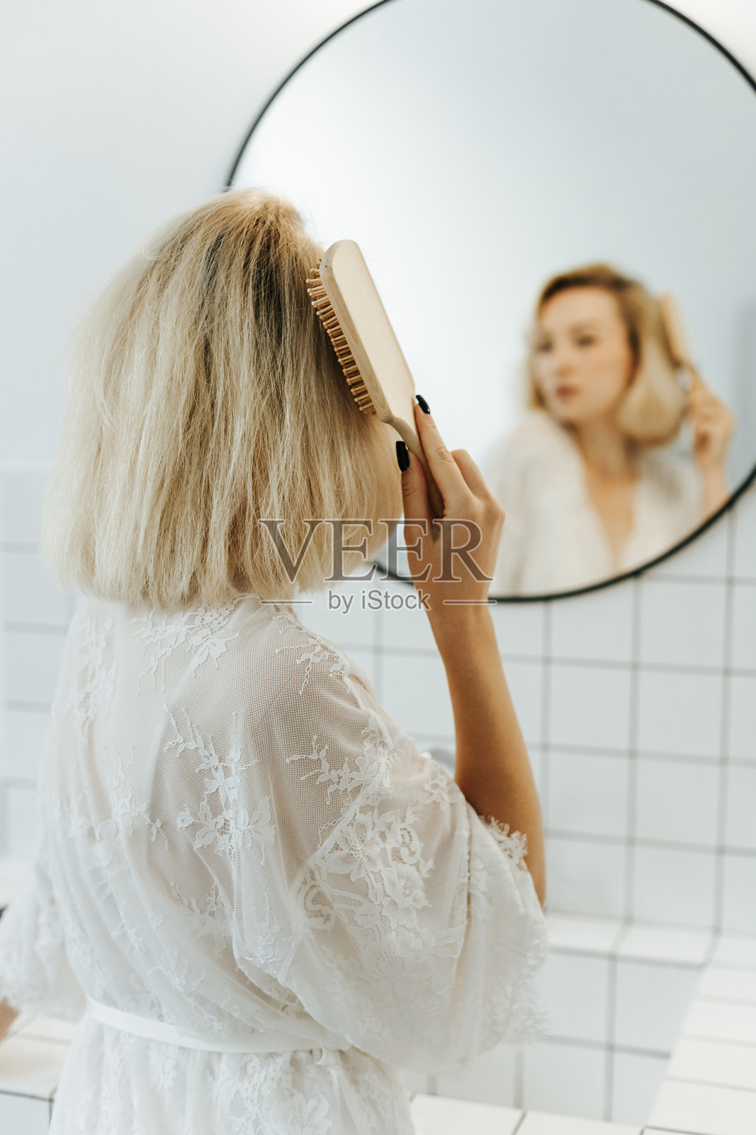 穿着白色背心的年轻女子在浴室里对着镜子梳头并微笑，这是她的背影。照片摄影图片