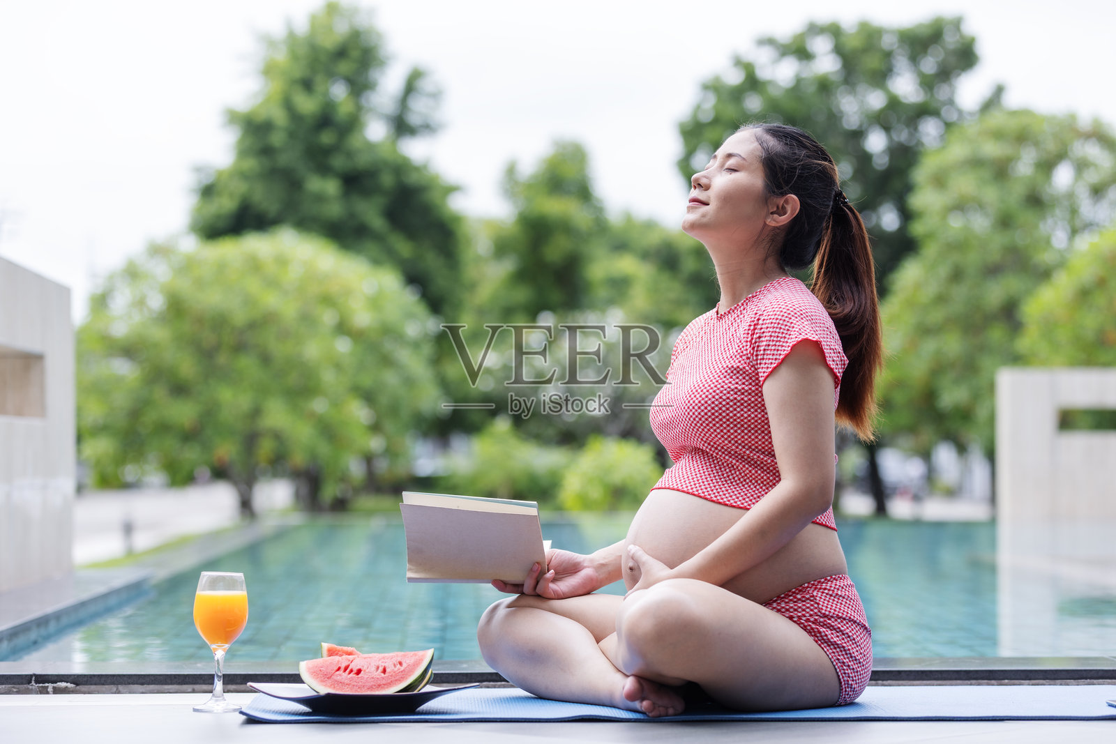 一位穿着红色格子连衣裙的孕妇平静地坐在泳池边，阅读书籍，享用新鲜的西瓜和果汁。照片摄影图片