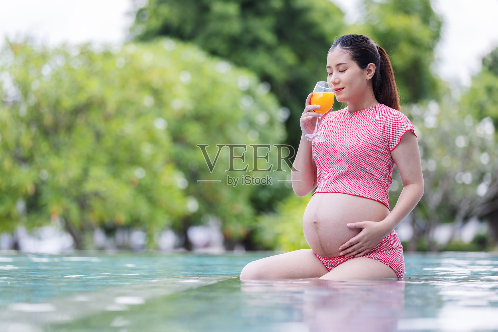 一位怀孕的女士穿着红色泳衣，在泳池中放松，手中拿着一杯橙汁。照片摄影图片