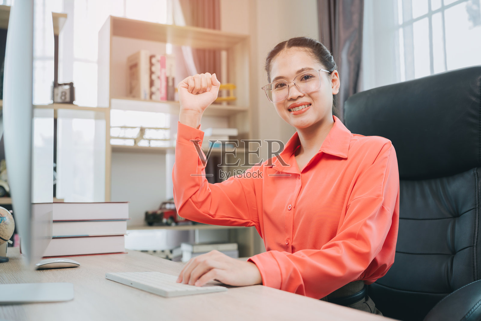 庆祝概念。年轻女子穿着橙色衬衫，在现代办公室里成功庆祝，同时使用电脑工作。照片摄影图片
